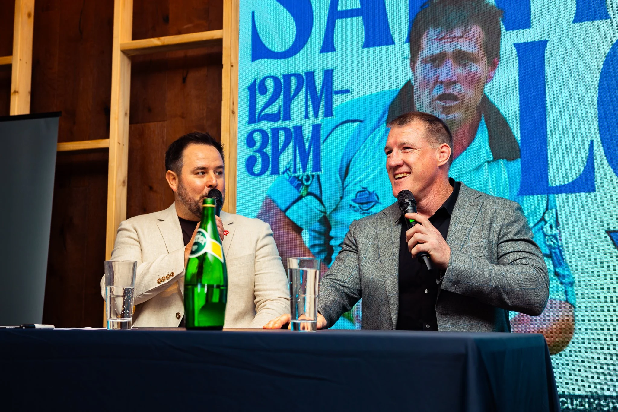 Two men are sitting at a table with microphones, glasses, and a bottle of water. They are speaking at an event, with a large screen behind them displaying their images and text. The man on the left has dark hair and a beard, wearing a light-colored b