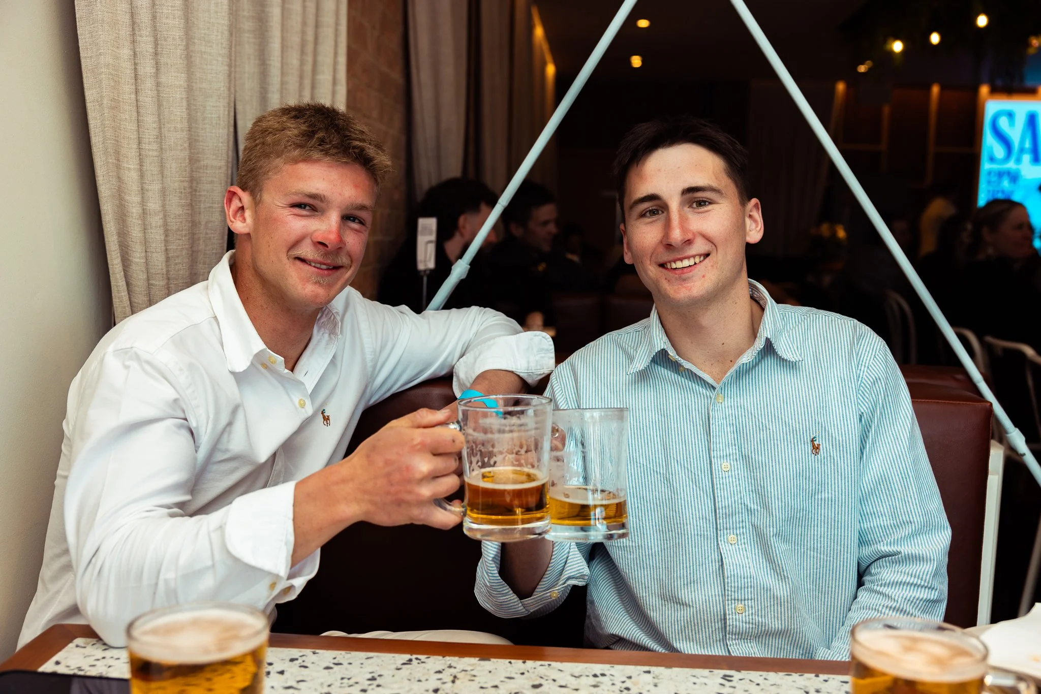 Two young men sitting at a restaurant table holding beer mugs and smiling at the camera.