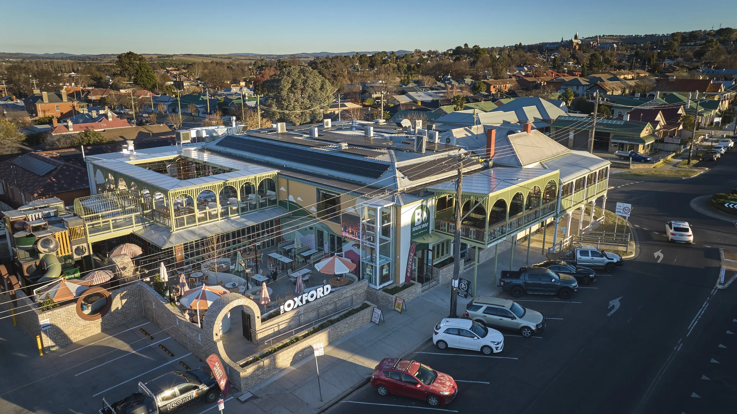 An aerial view of a shopping complex with outdoor seating, a parking lot filled with cars, and a surrounding neighborhood with houses and trees. The complex has a large sign that reads 'Oxford' and features solar panels on its roof.