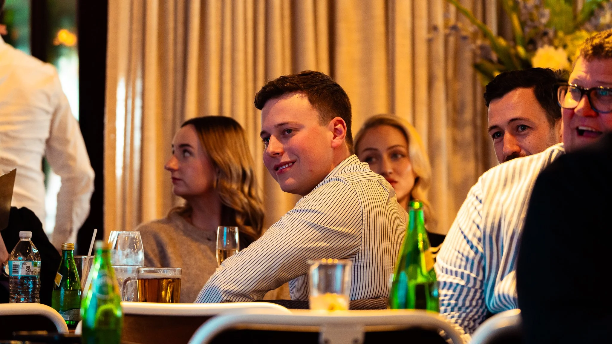 People attending a formal event sit at a table with drinks and bottles, listening attentively