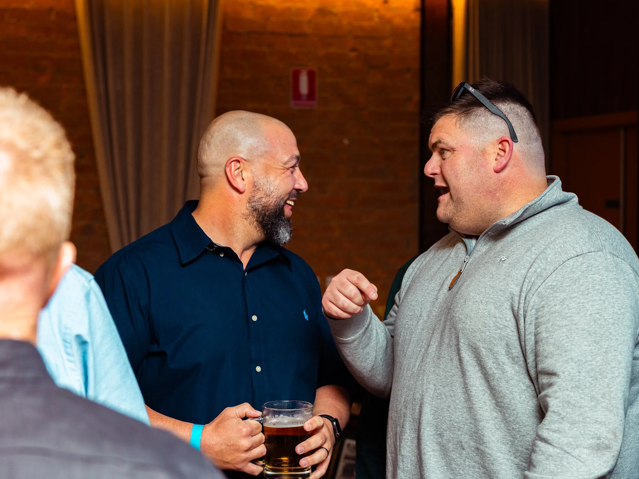 Two men are engaged in conversation, with one holding a glass of beer. The setting appears to be indoors with warm lighting and wooden walls.