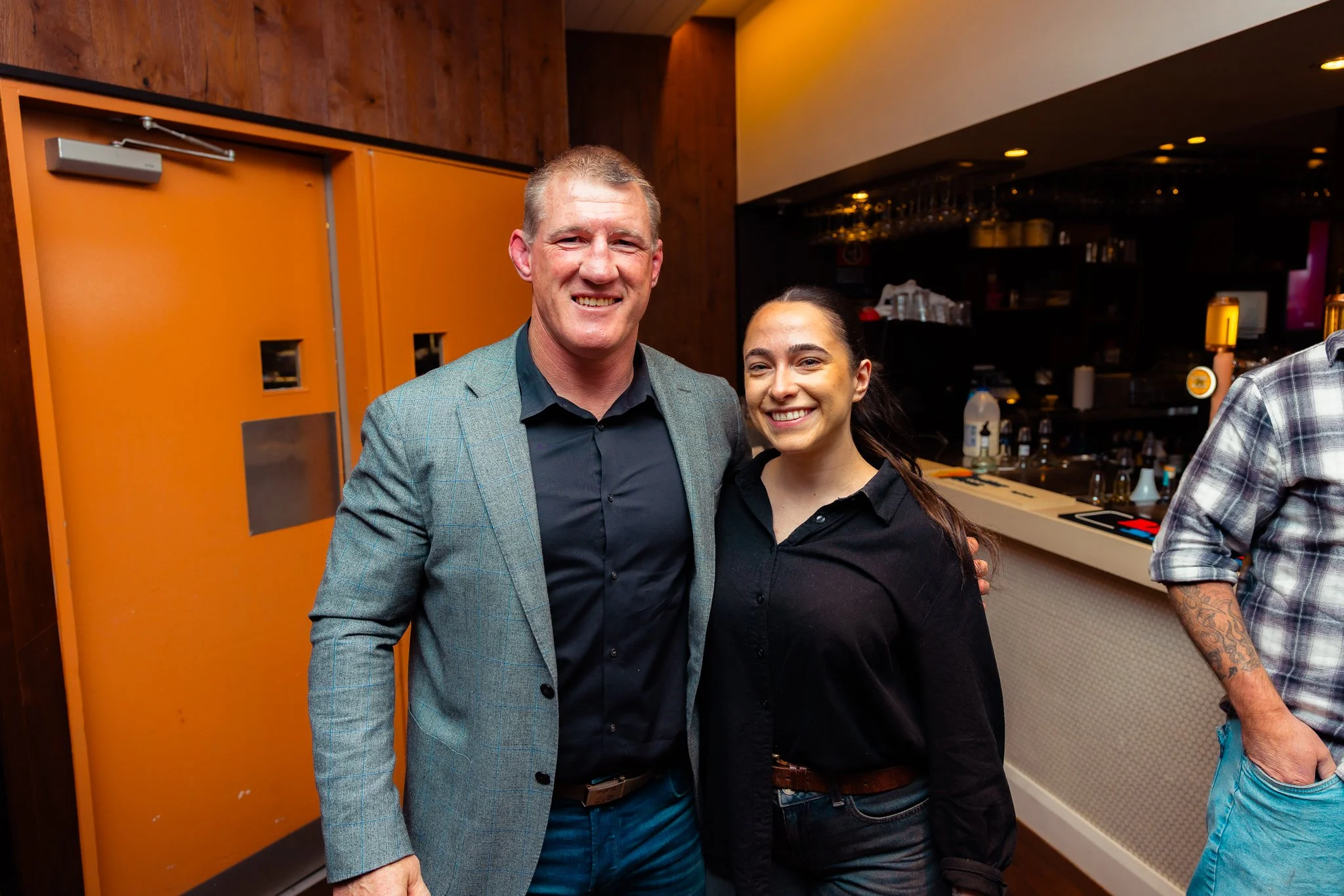 A man in a green blazer and a woman in a black shirt standing close together and smiling at the camera. The background shows a bar area and a bright orange door.