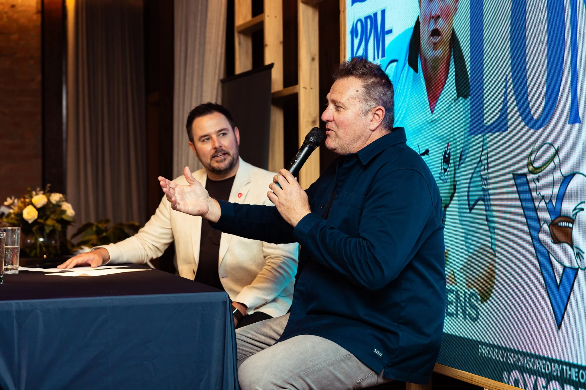 Two men sitting at a table during a panel discussion or interview. The man on the right is speaking into a microphone, wearing a dark shirt with gray pants. The man on the left is listening, dressed in a light-colored blazer and dark shirt. There is 