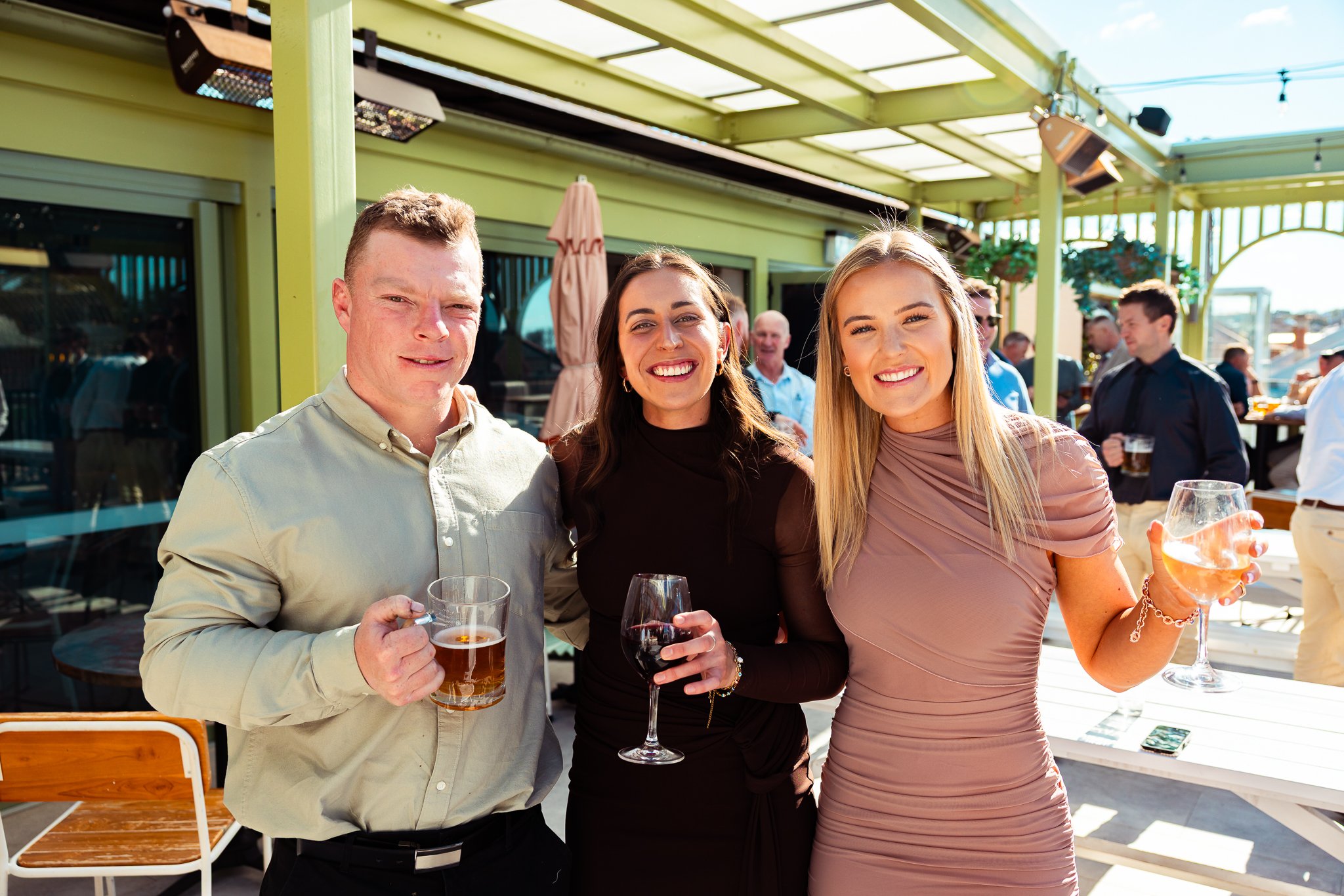 Three friends enjoying drinks at an outdoor event or patio, smiling at the camera with a green canopy overhead, other people in the background, and holding wine and beer glasses.