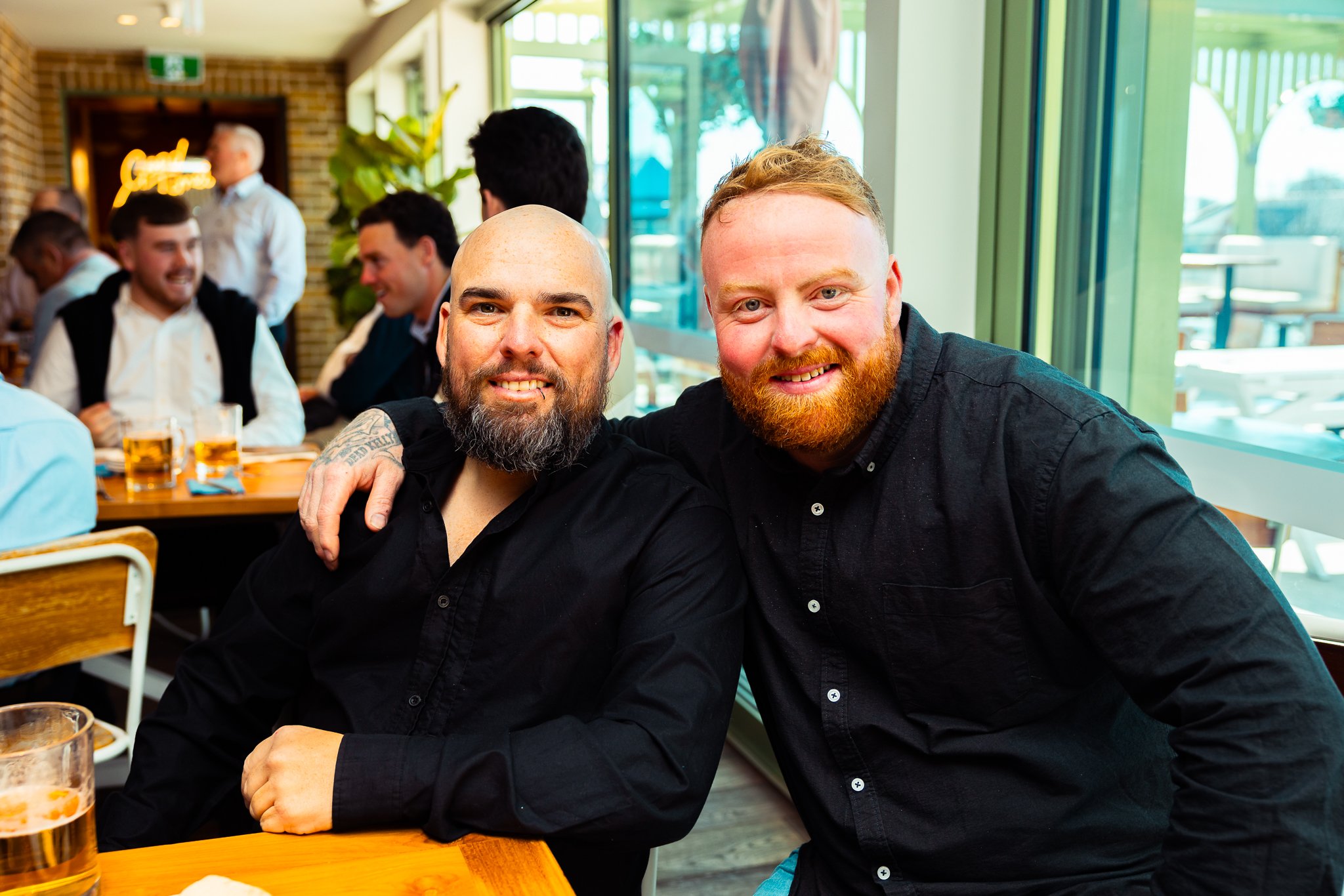 Two men with beards smiling at the camera in a restaurant. The man on the left is bald with a beard, and the man on the right has red hair and a red beard. They are sitting at a table with drinks and are surrounded by other people in the background, 
