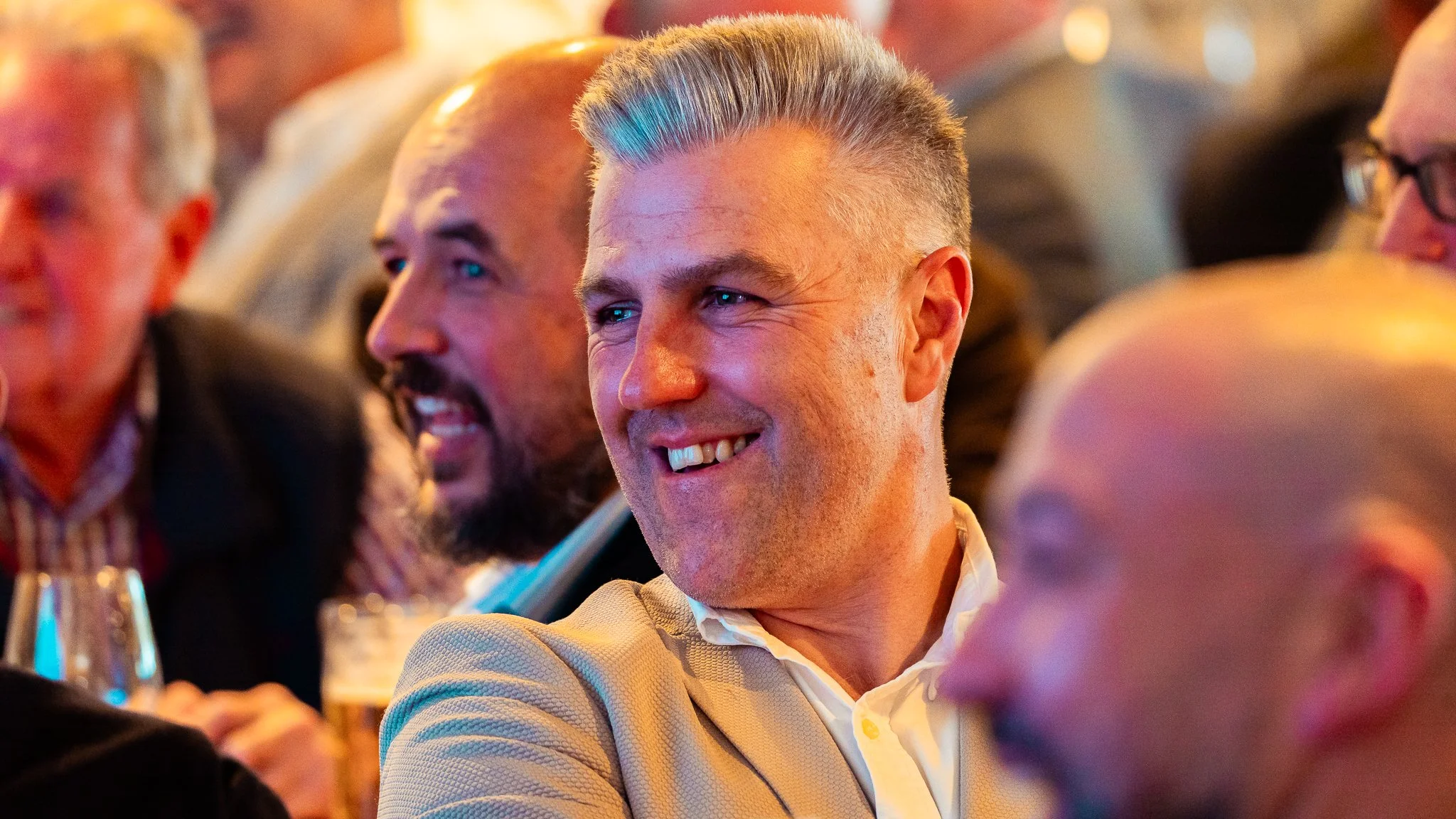 A group of people at a social gathering, smiling and engaging in conversation. The focus is on a smiling man with gray hair, wearing a light-colored blazer and white shirt.