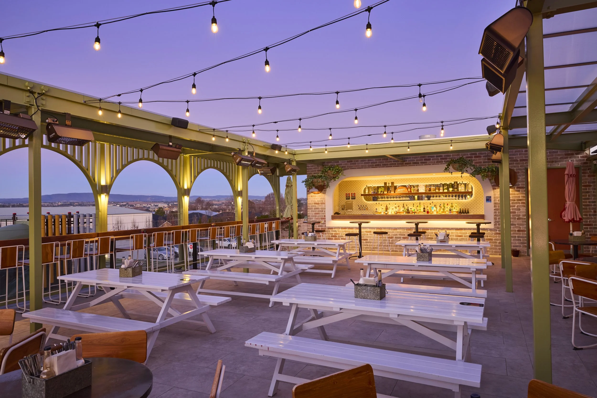 Outdoor rooftop bar or restaurant with string lights hanging in the sky, bar area with shelves of bottles, white picnic tables and chairs, a few potted plants, and a view of a city or suburban area in the distance during twilight.