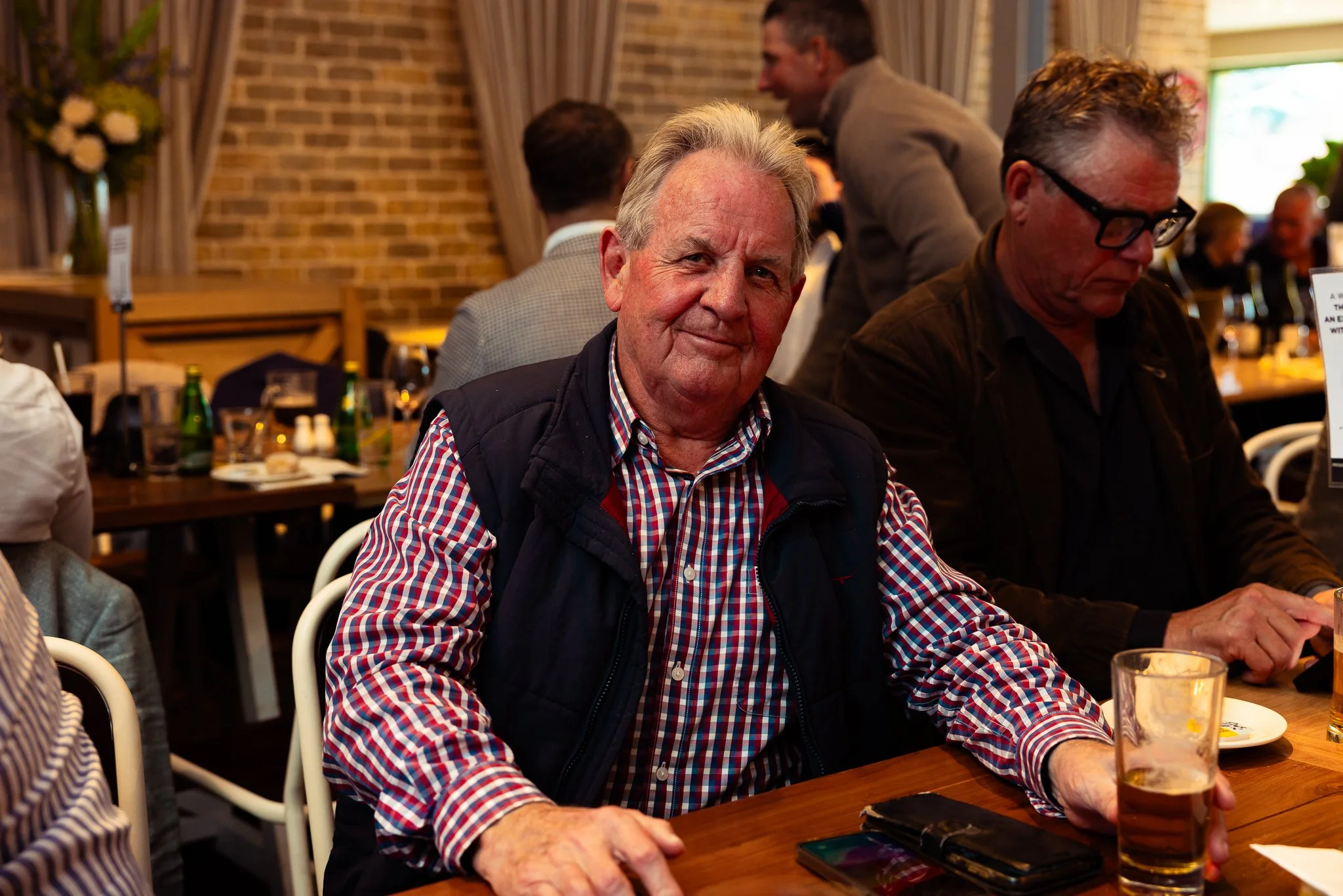 Older man with gray hair wearing a checkered shirt and a dark vest sitting at a table in a restaurant, smiling at the camera. Other people are visible in the background, some engaged in conversation, with a brick wall and floral decoration in the res