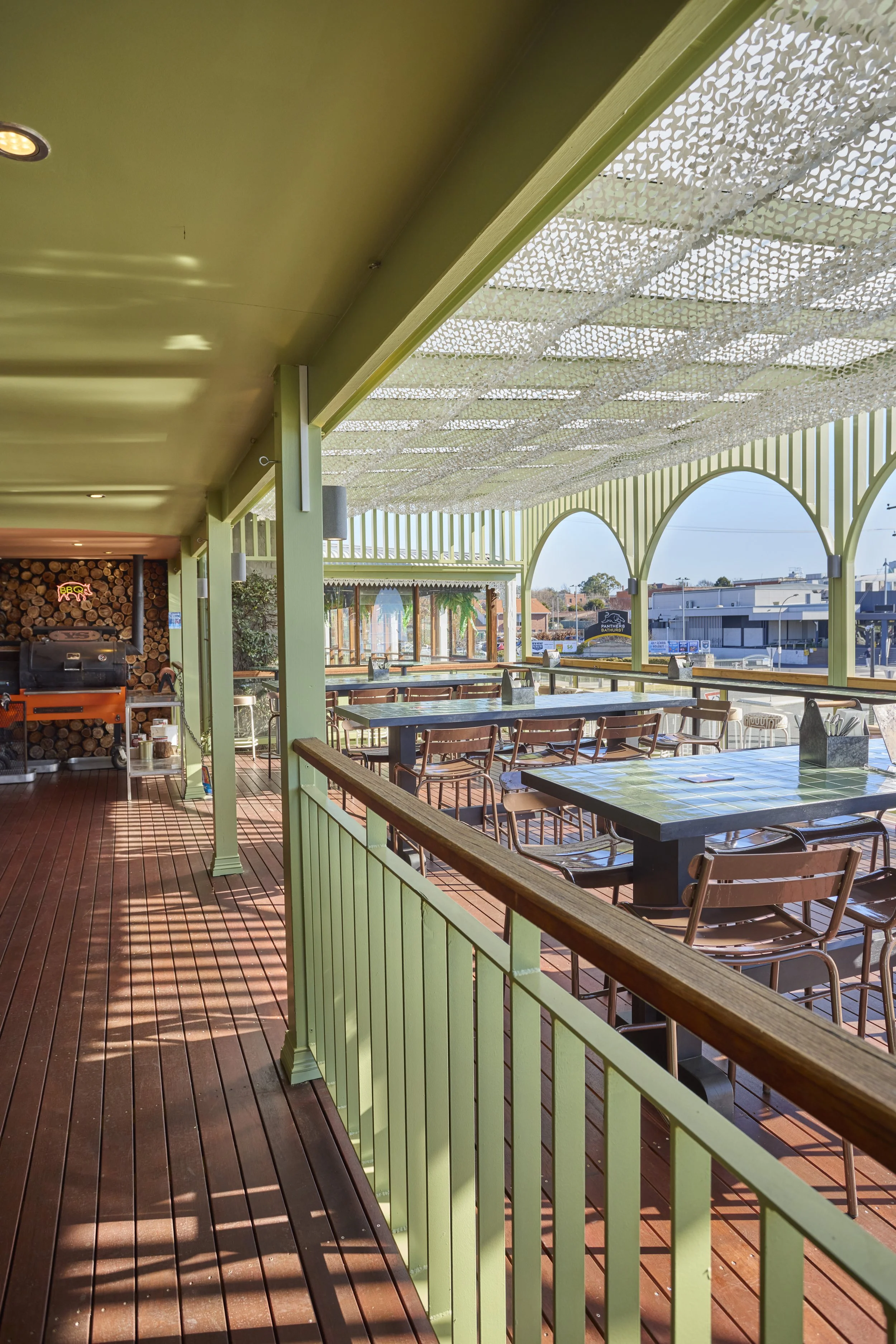 Outdoor patio area with green painted wooden railings, tables, chairs, and a decorative overhang with a perforated design providing partial shade. In the background, buildings and a street are visible.