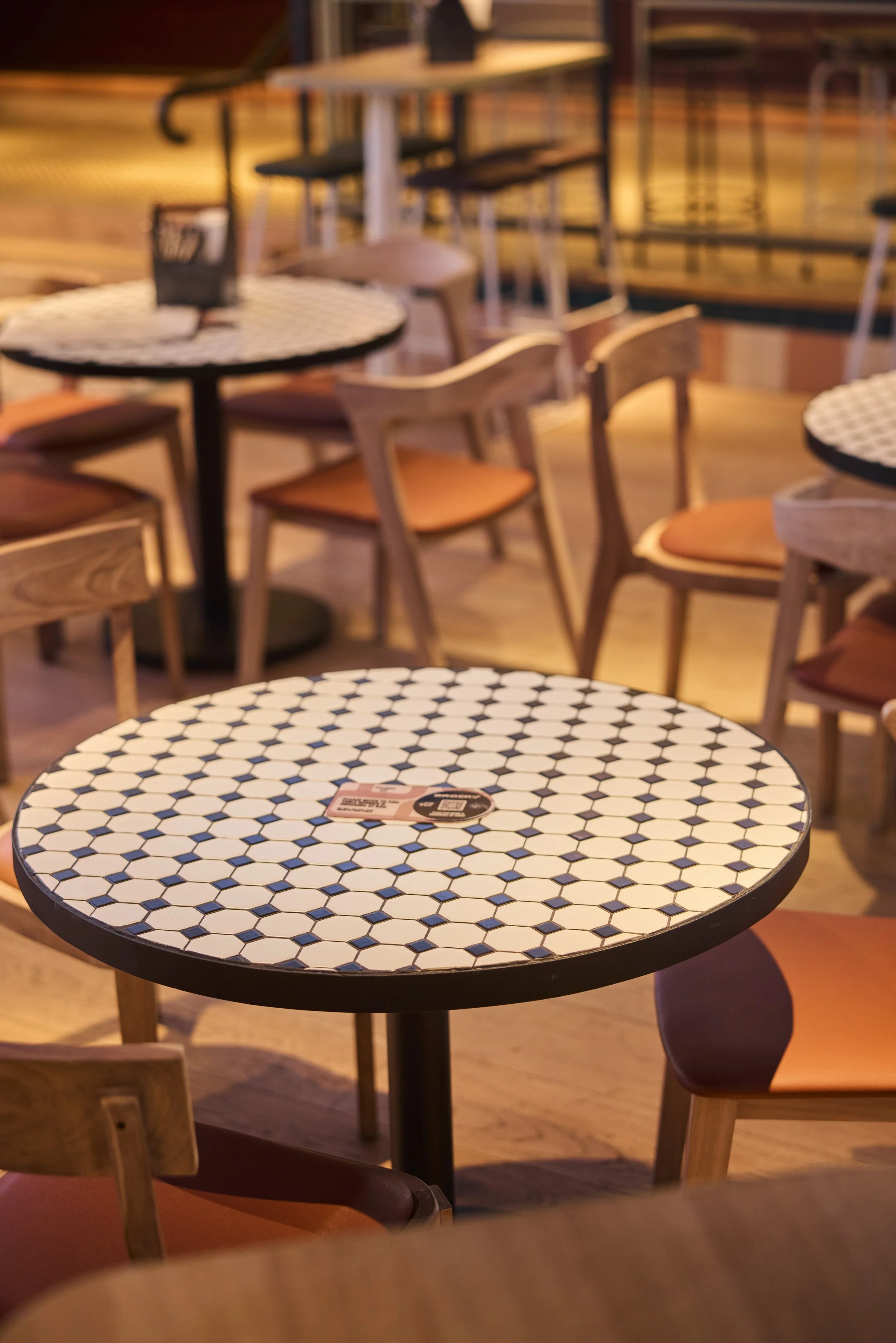 Blurry interior of a restaurant with round tables and wooden chairs, one with a black and white hexagonal patterned top in the foreground.