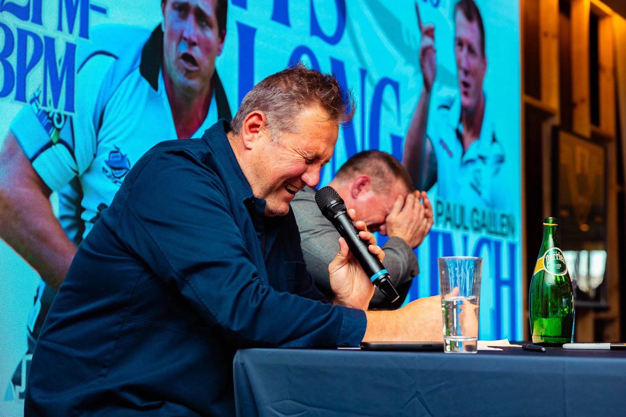 A man with gray hair and a dark shirt laughing and holding a microphone at a panel or event, with two other men beside him covering their faces, behind a blue backdrop with text and images.