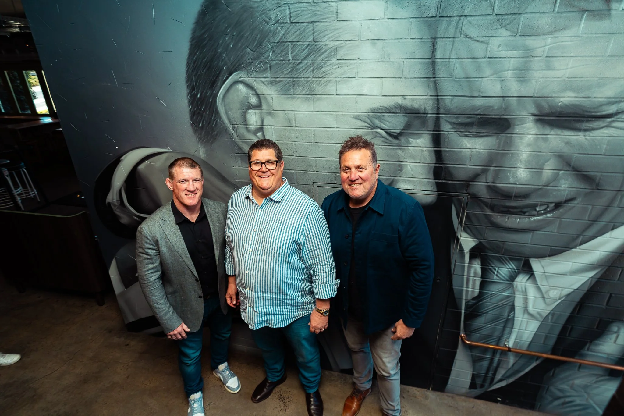 Three men standing in front of a mural of a man wearing glasses and headphones, with a brick wall in the background.