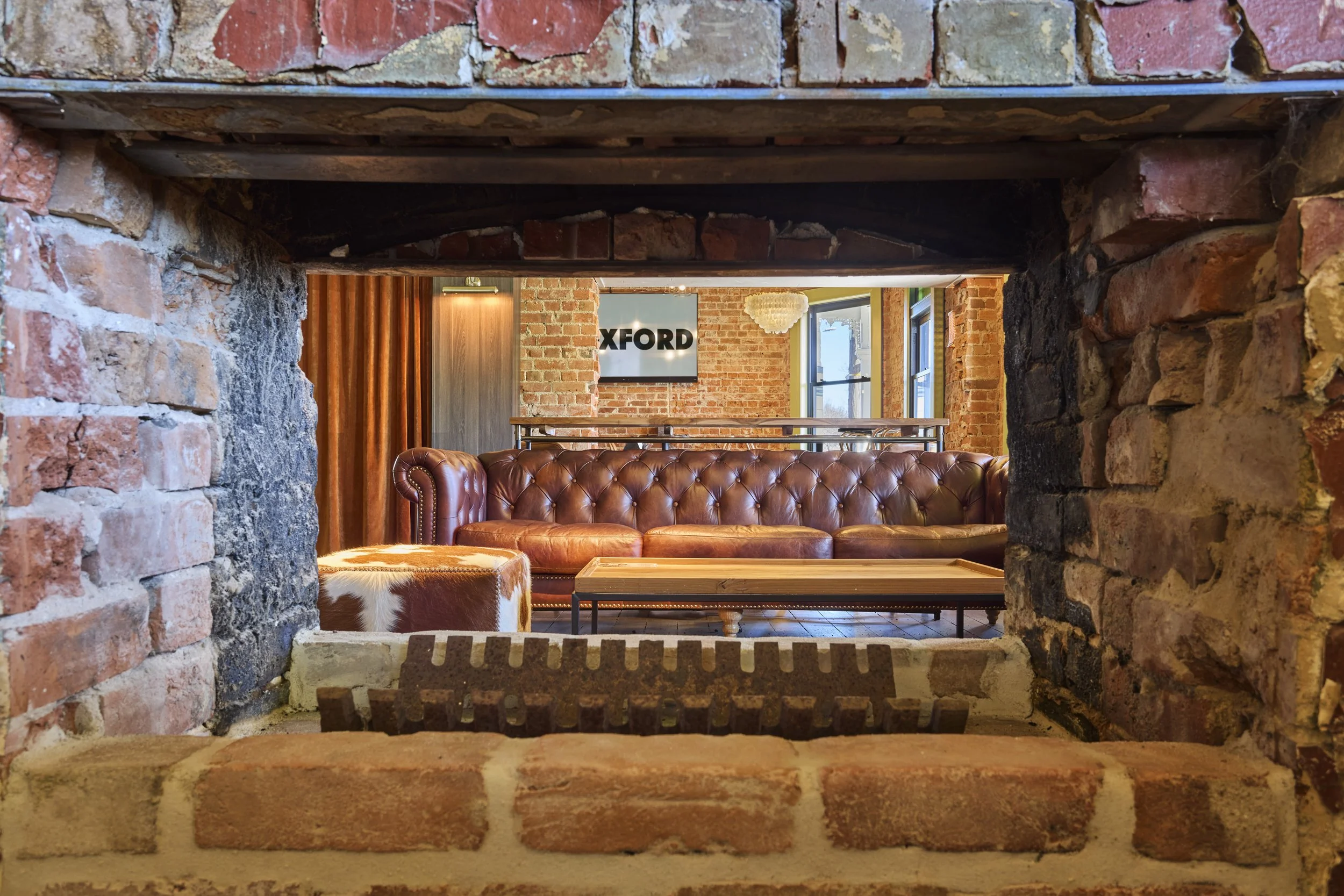 View through a brick fireplace opening into a modern industrial living room with exposed brick walls, large windows, and a leather tufted sofa.