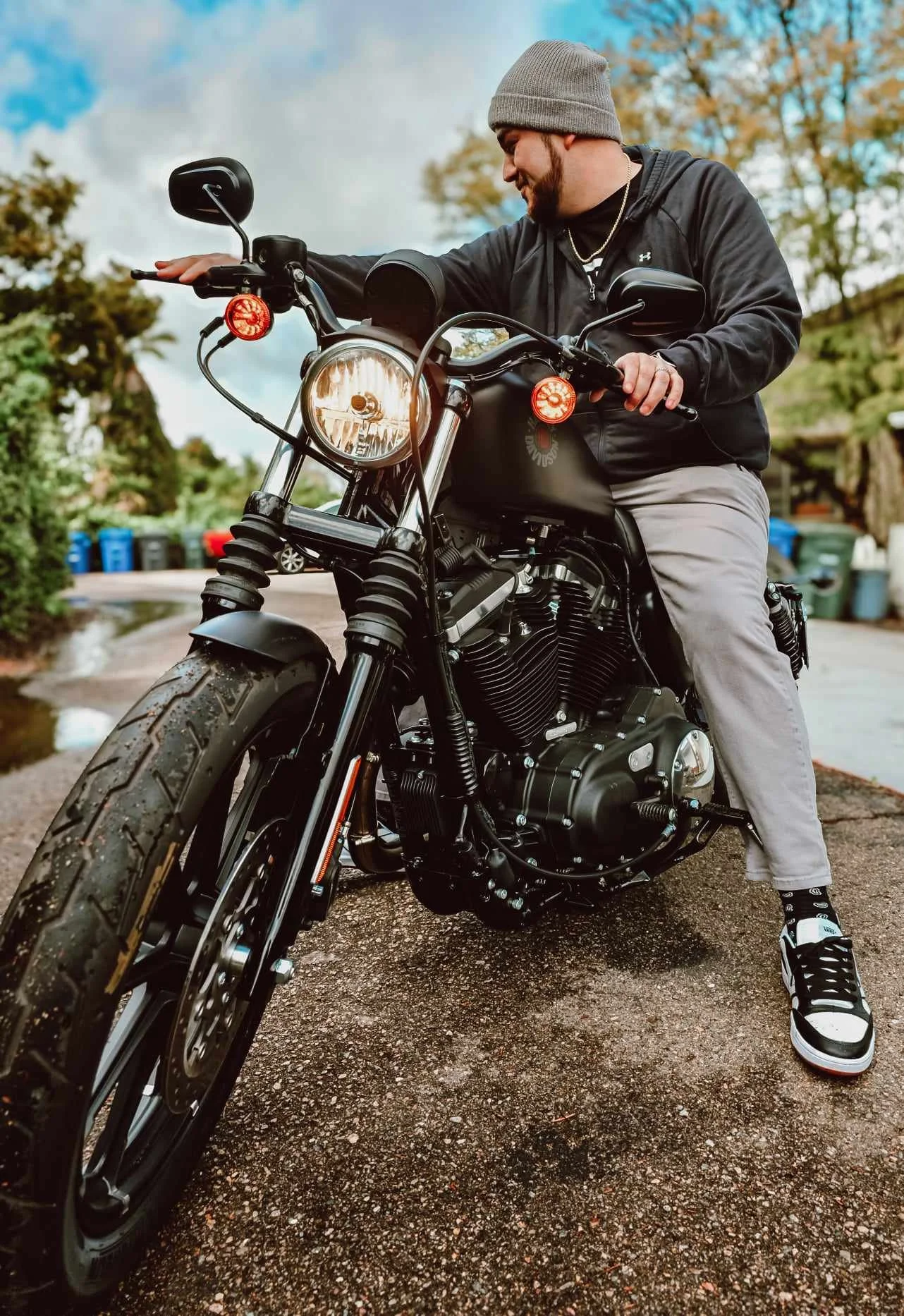 A man wearing a gray beanie, black jacket, and light pants sits on a black motorcycle outdoors on a cloudy day, with trees and recycling bins in the background.