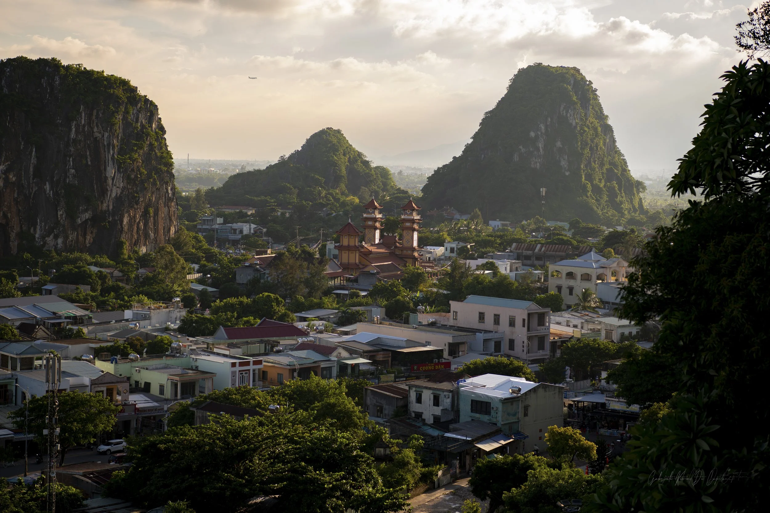 A cityscape with a cluster of houses and buildings at the base of large, green, rocky mountains (Vietnam's Marble Mountains), with an airplane flying in the sky above.