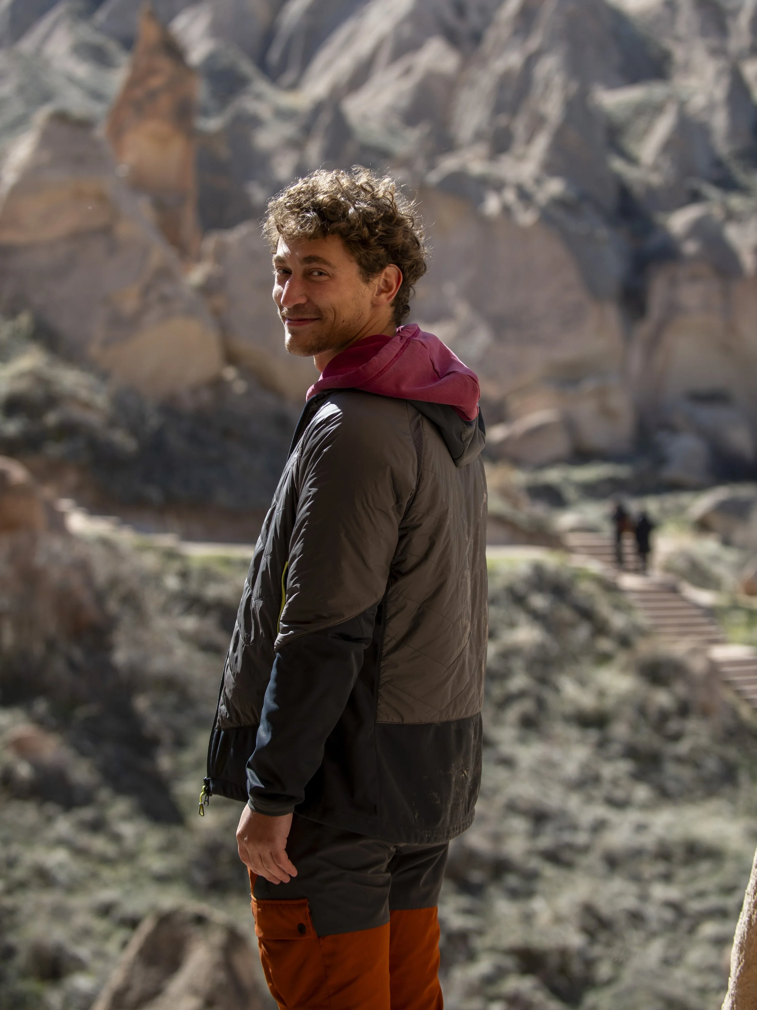 A man with curly hair smiling and looking over his shoulder, wearing a black jacket with a red hood and orange pants, standing outdoors with a rocky background.