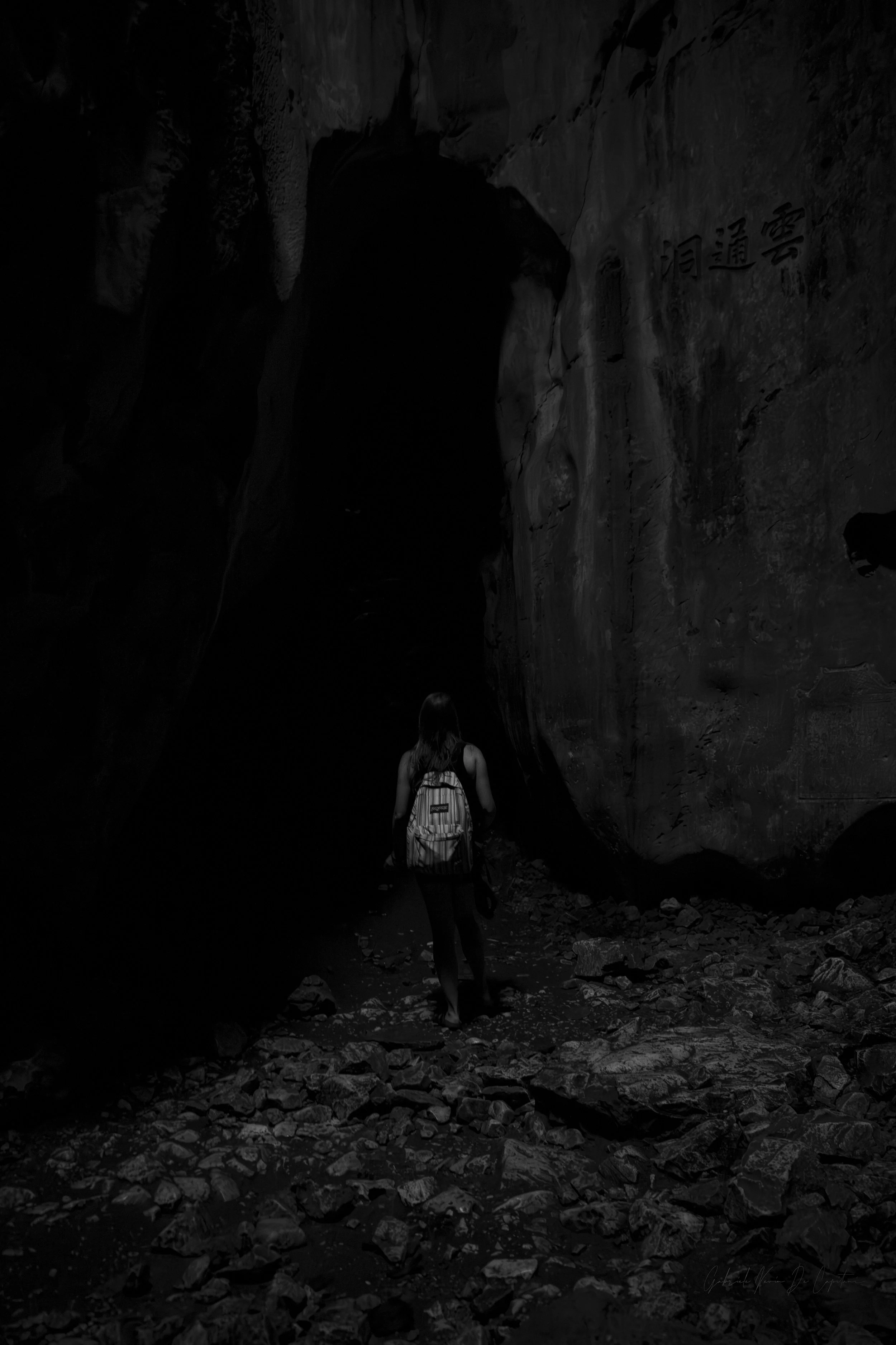 A person with a backpack walking through a dark, rocky cave entrance with Chinese characters carved on the wall.