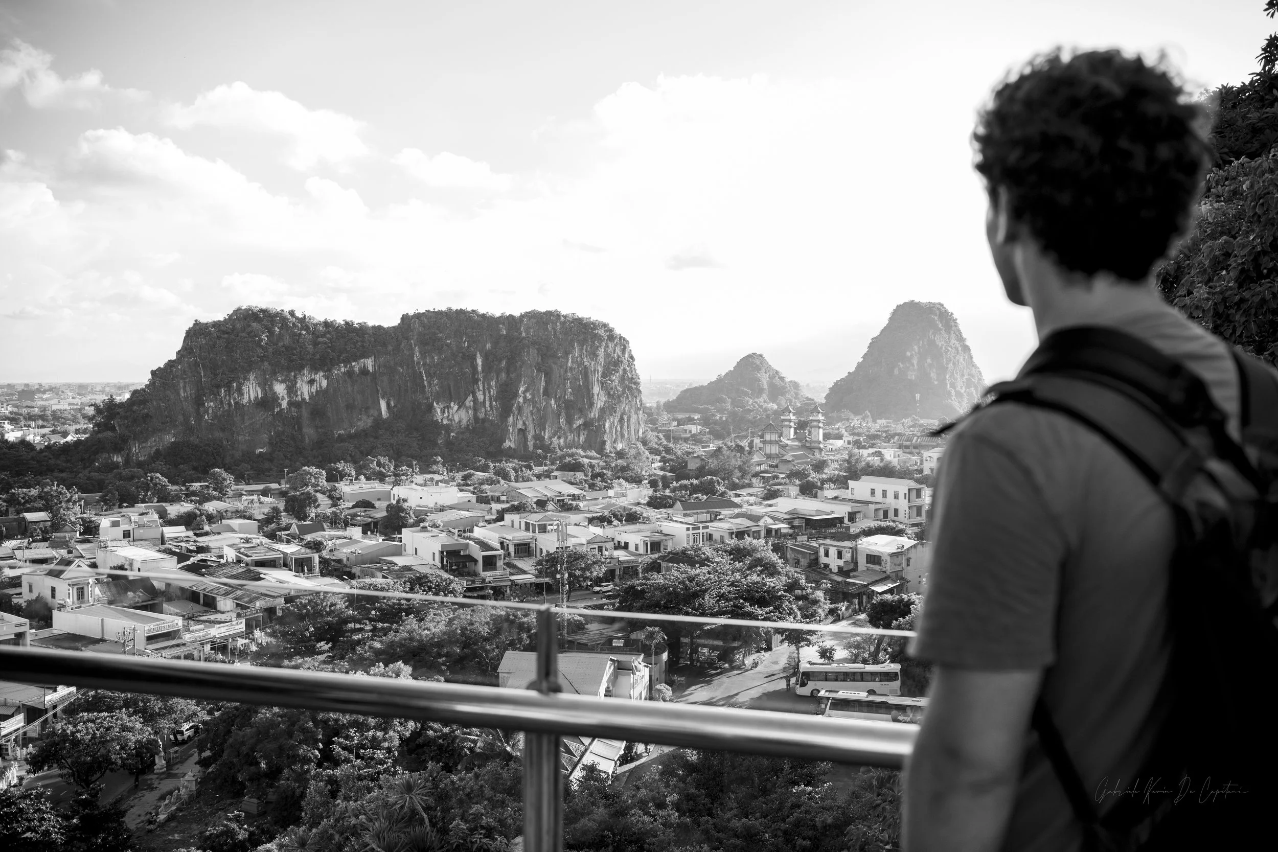 A person with short hair and a backpack looking out from a balcony over a city with Vietnam's Marble Mountains in the background.