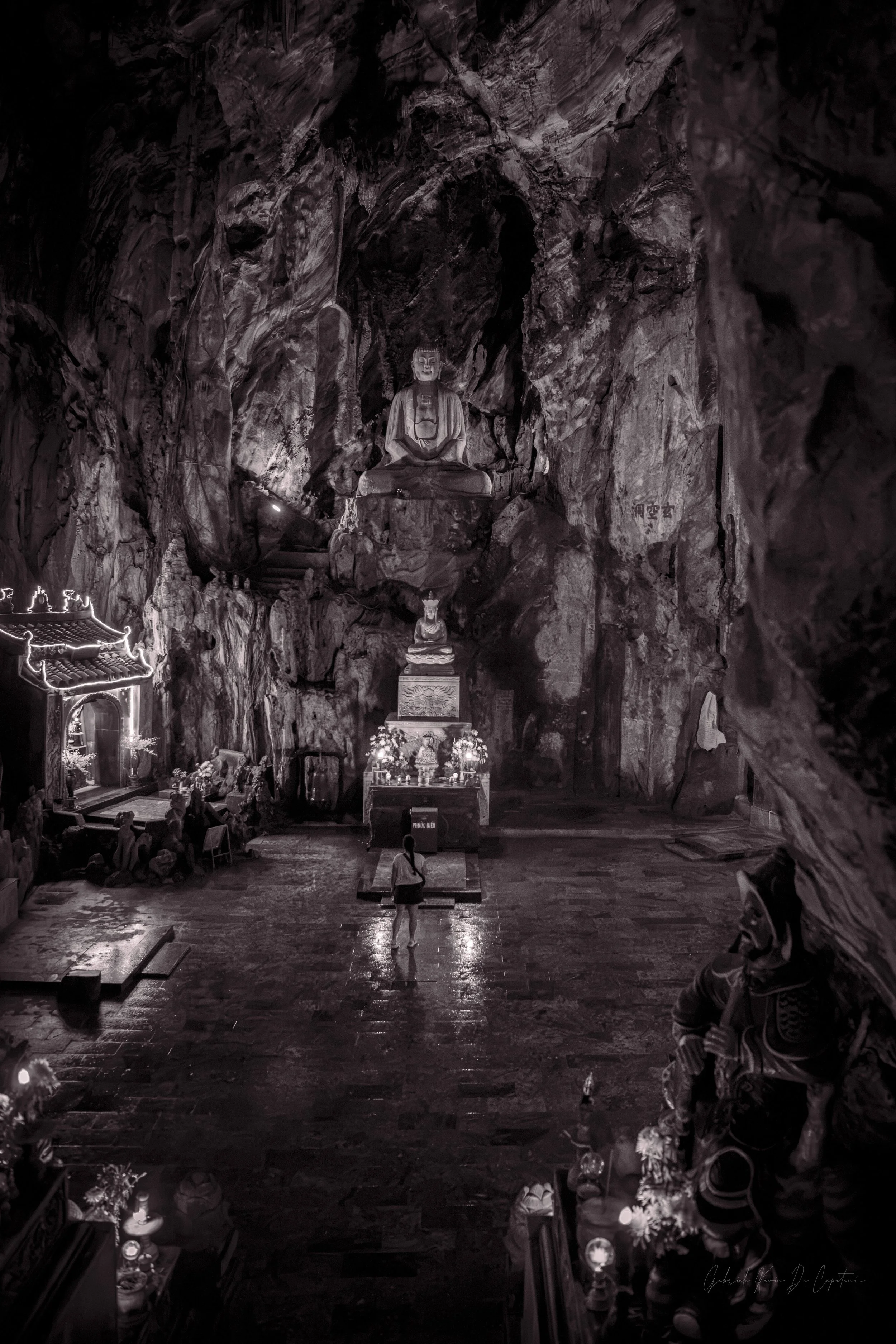 Inside a large, dark cave with a carved Buddhist statue at the back, lit by candles and spotlights. The floor is wet and reflective. One young woman is standing admiring it, within the Buddhist temple carved inside Vietnam's Marble Mountains.