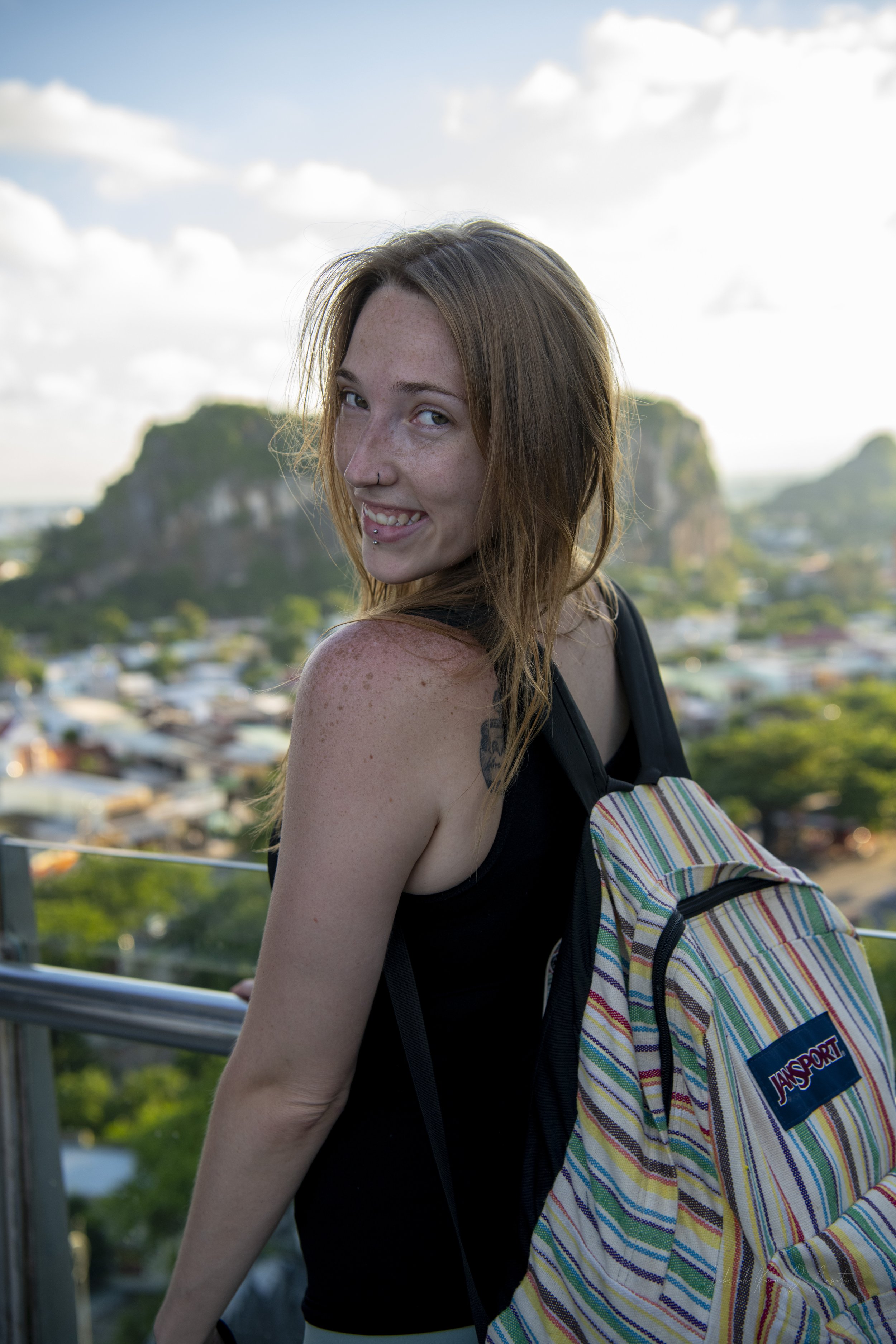 A young woman with red hair and freckles, smiling and sticking out her tongue, carries a colorful striped backpack on her shoulder, in an outdoor setting with a cityscape and a hill in the background.