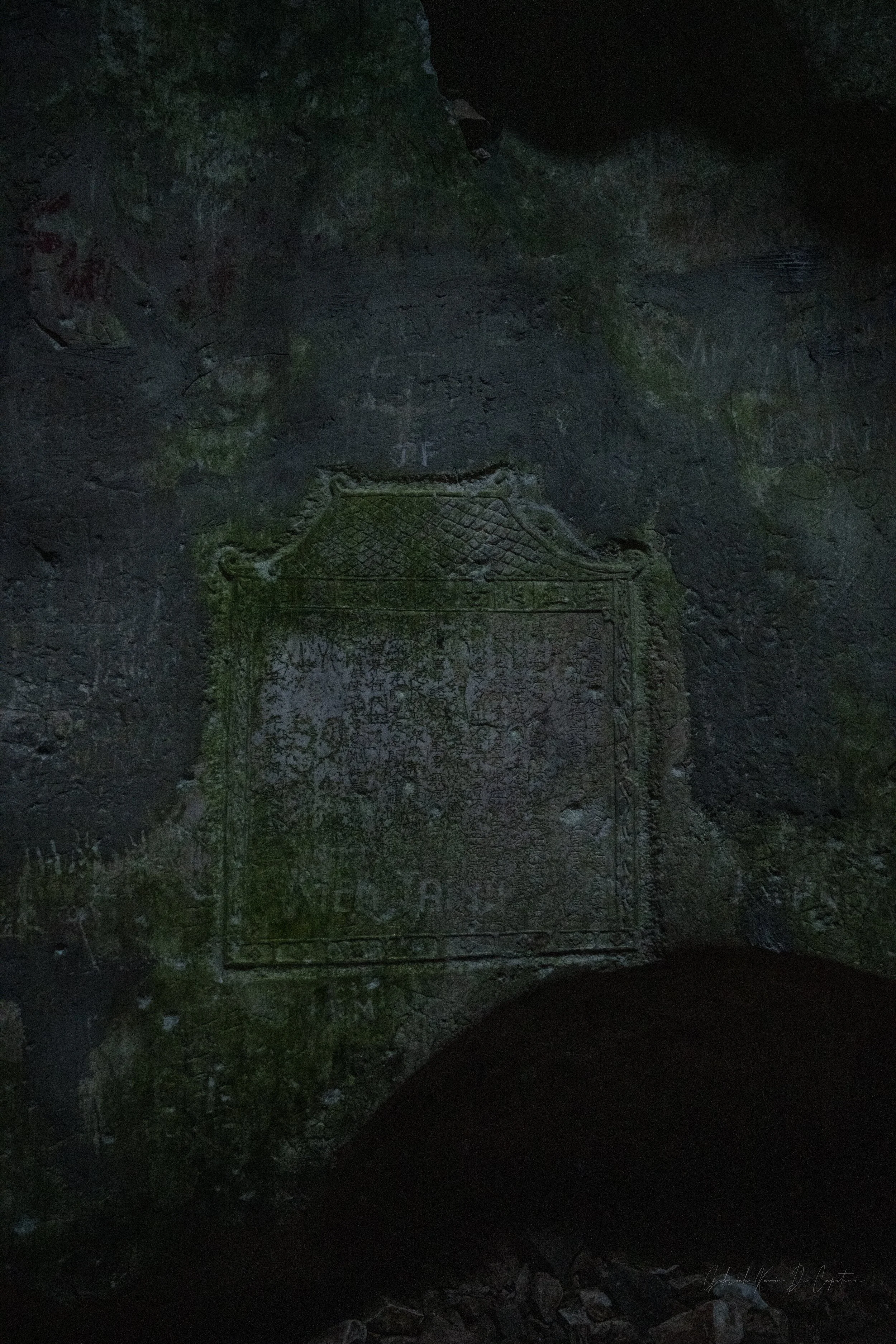 A dark, moss-covered rock wall with engraved Chinese characters and decorative carvings, illuminated faintly to reveal inscriptions.