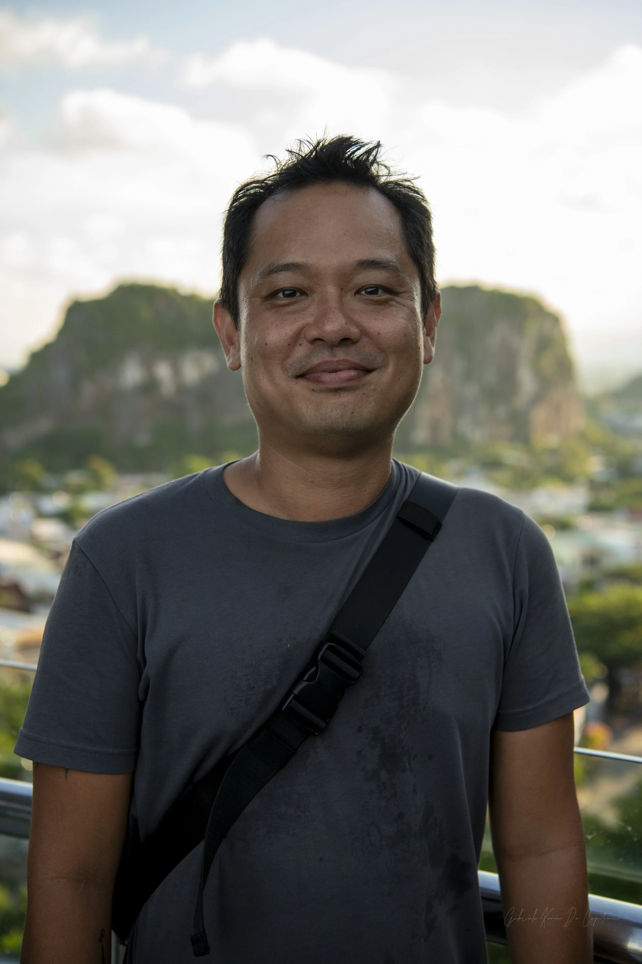 A man with short dark hair and a slight smile stands outdoors with a mountainous landscape and partly cloudy sky in the background.