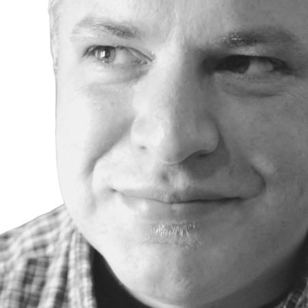 Black and white image of a person smiling, wearing a checkered shirt, with a close-up on the face.