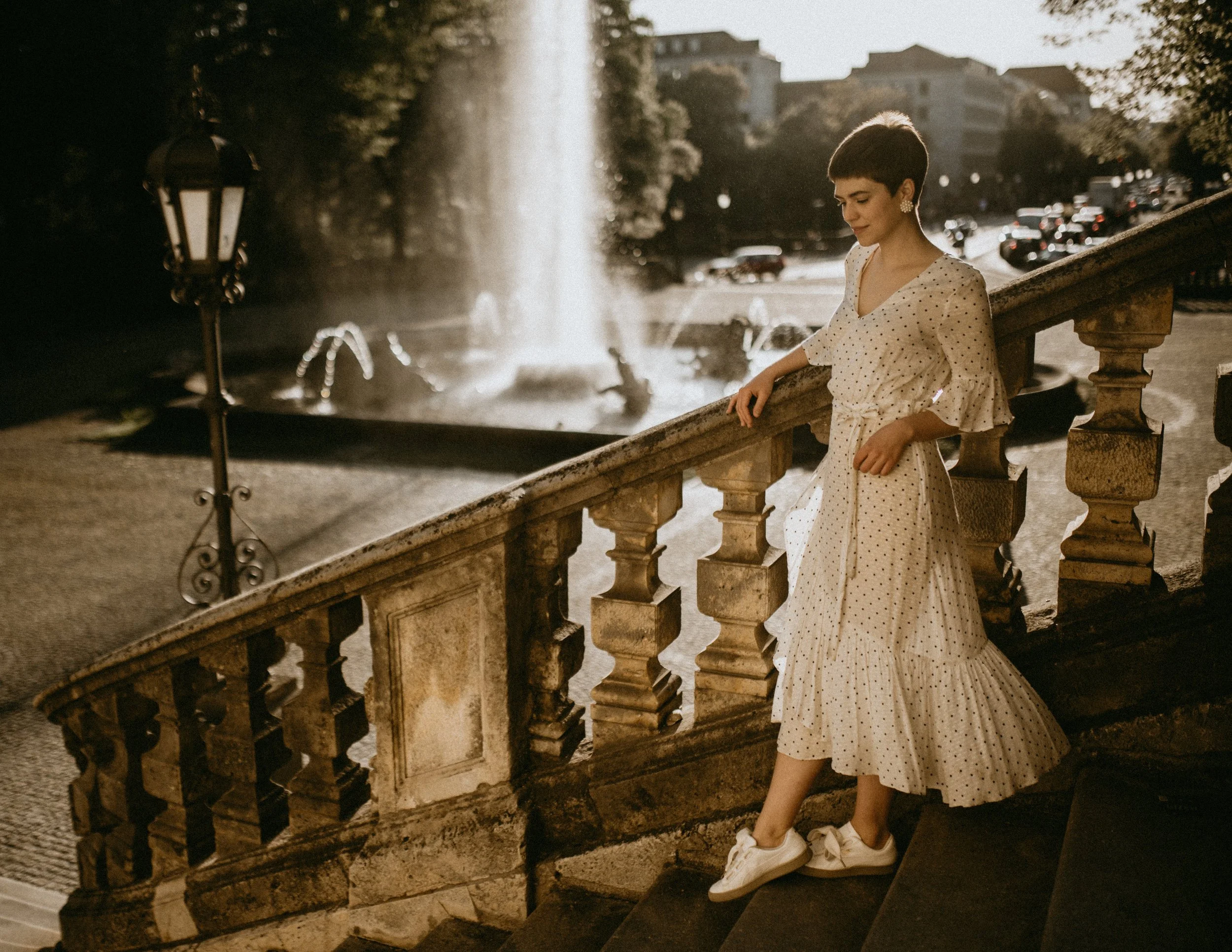 Woman in white polka dot dress standing on stone staircase next to a fountain in an urban park setting during sunset.