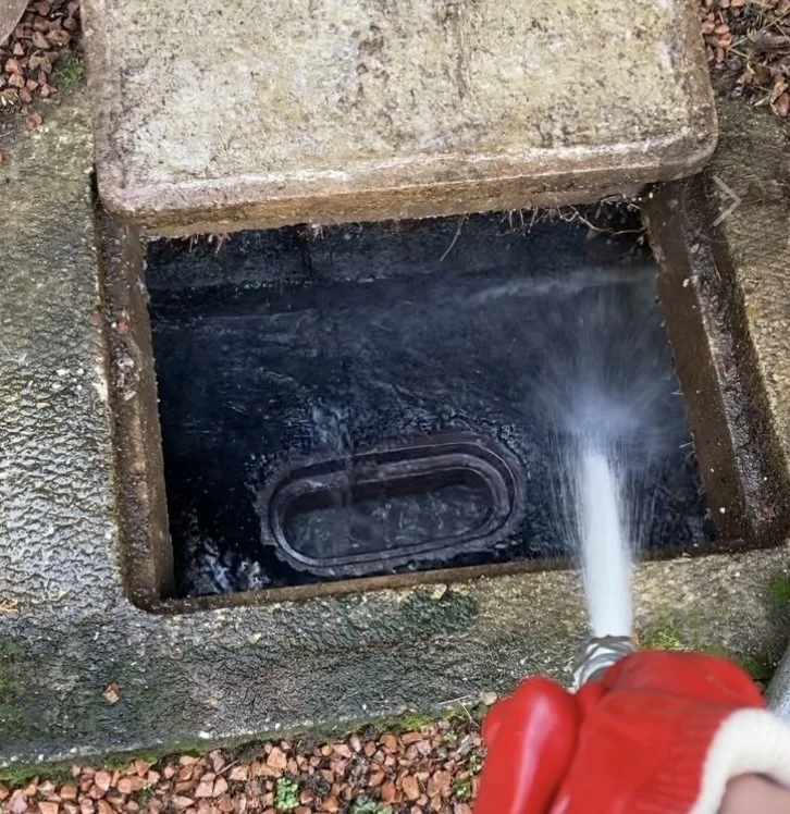 A person in red gloves is using a hose to spray water into a storm drain or sewer opening on a concrete surface outside.