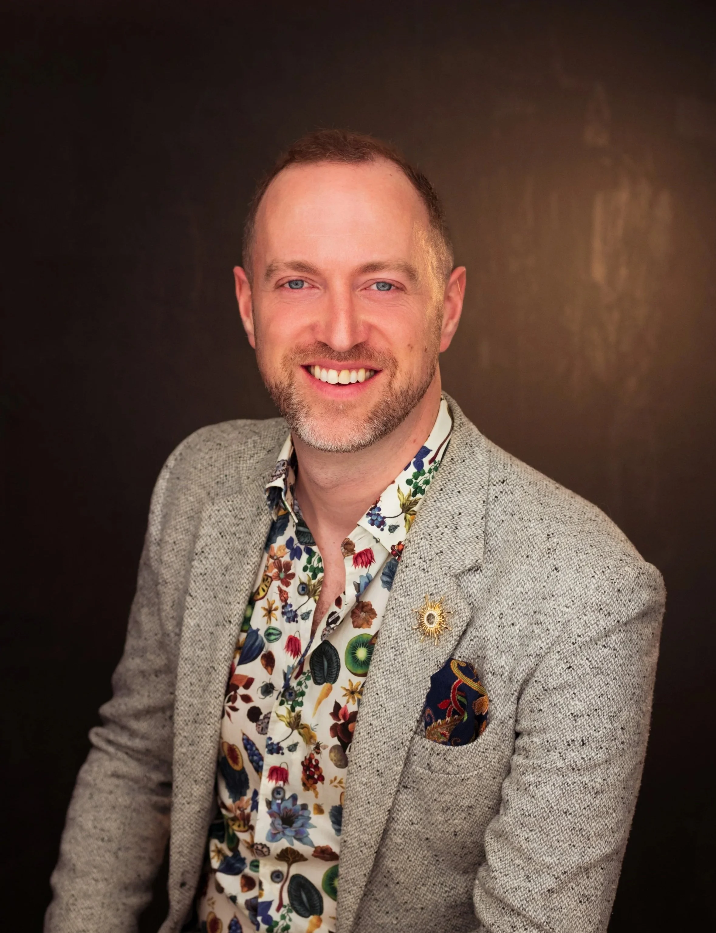 A man with short light brown hair, blue eyes, and a beard, smiling and wearing a gray blazer, a floral shirt, and a decorative pin and pocket square, against a dark background.