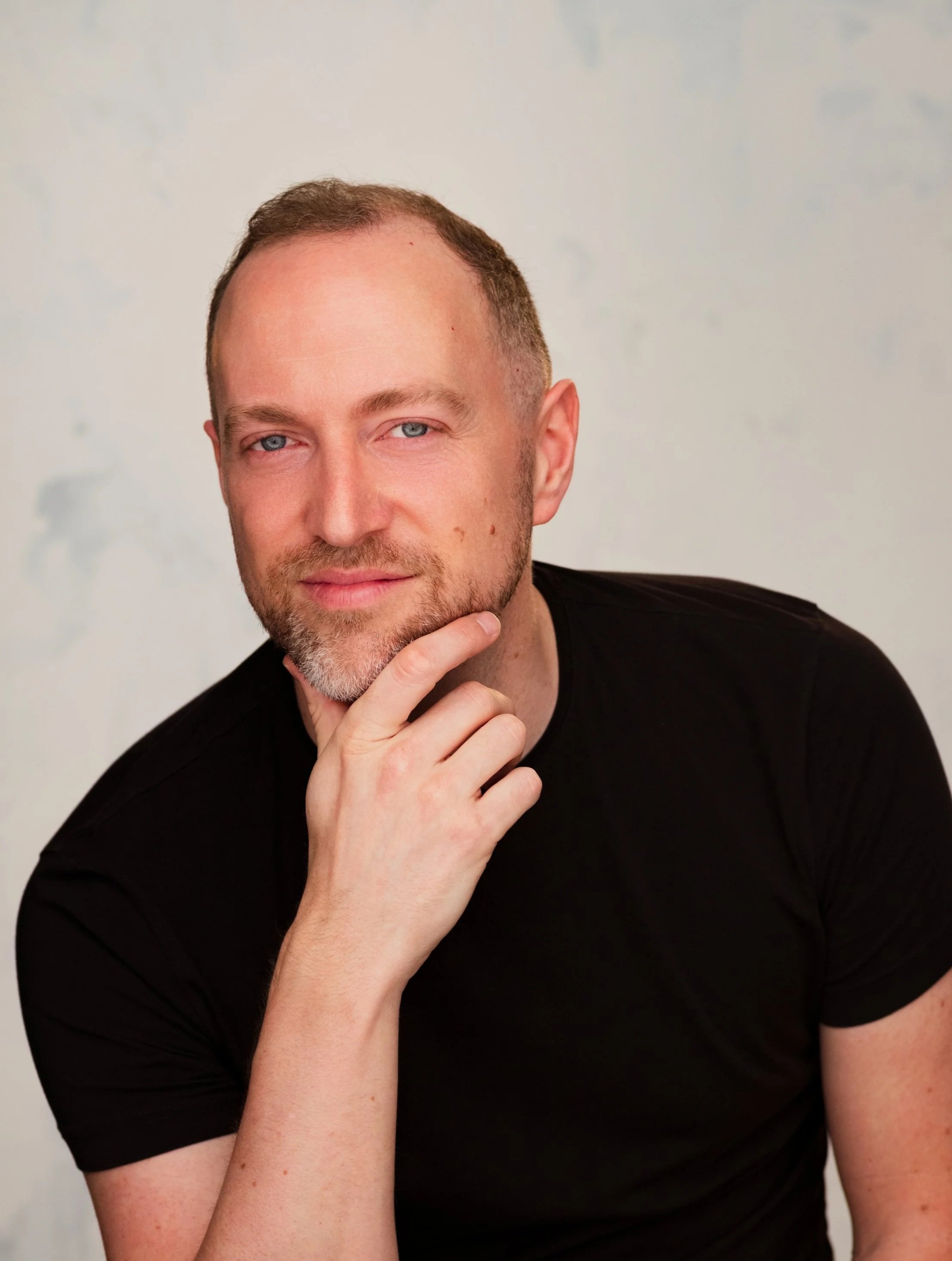 Close-up of a man with short brown hair and a beard, wearing a black shirt, with his hand on his chin, posing thoughtfully against a plain background.