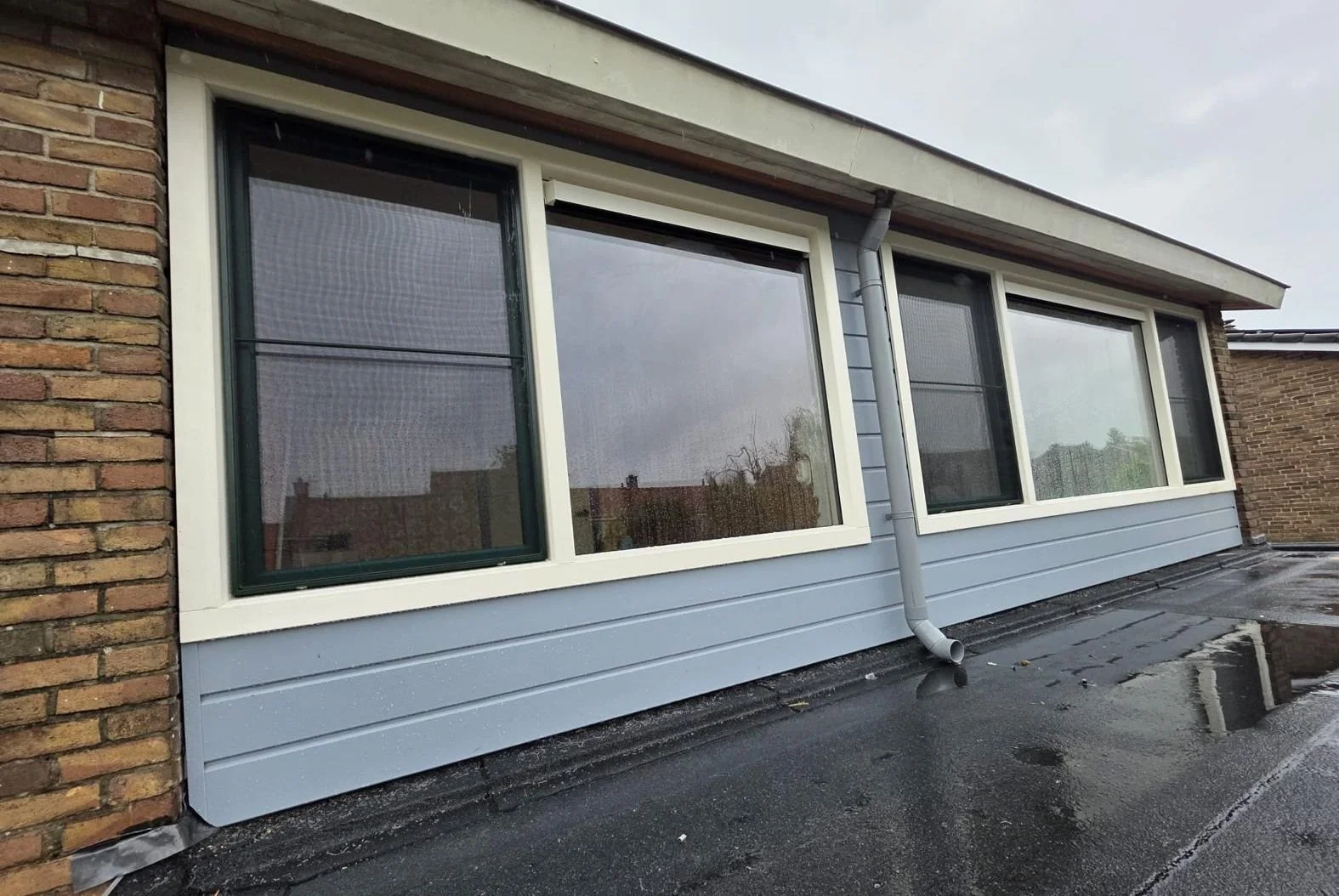 Uitzicht op een zolderraam met drie grote ramen, een windafvoerpijp en een zwart asfaltpad eronder, in een woning met blauwe gevelbekleding en een bakstenen muur, tijdens een regenachtige dag.