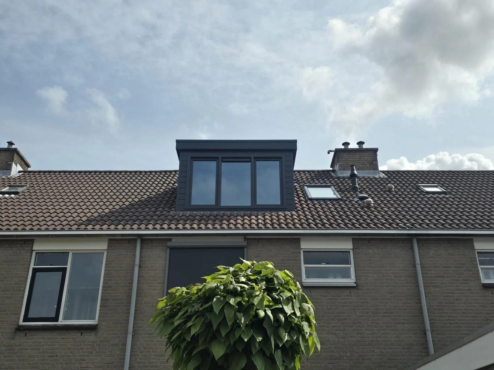 Een huis met een dak met nokvorsten en verschillende ramen, waaronder een grote glazen dakkapel en enkele kleine ramen, onder een bewolkte hemel.