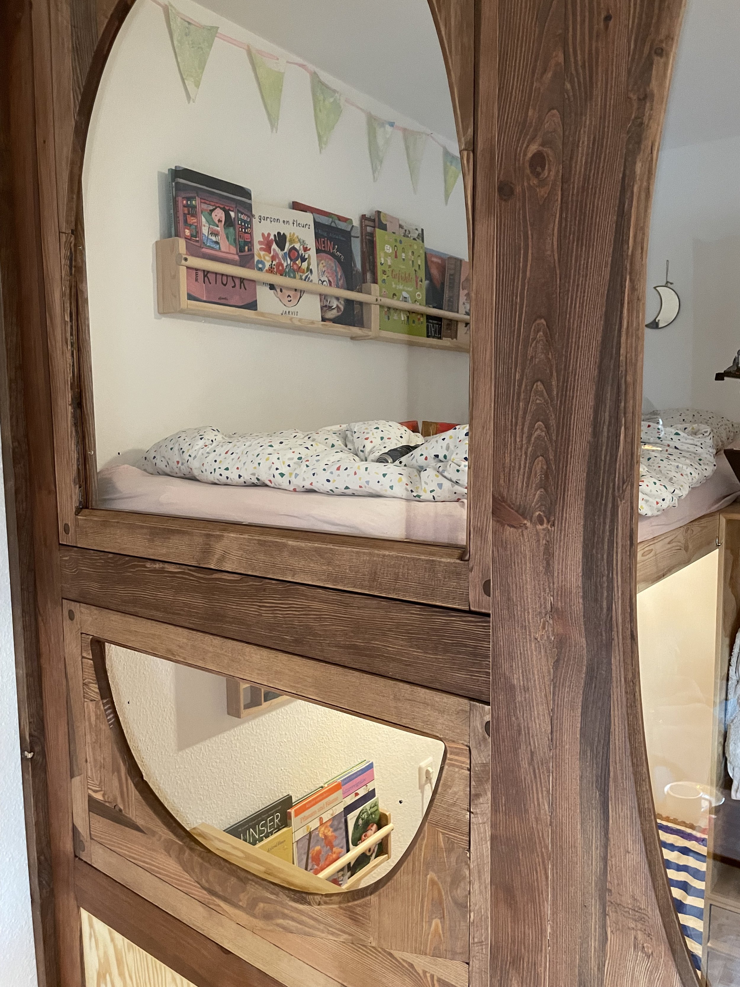 Ein Hochbett aus Holz mit einer Matratze und buntem Bettzeug, Blick durch das offene Fenster. Im Hintergrund sind Bücherregale und eine Wand mit Wanddekorationen zu sehen.