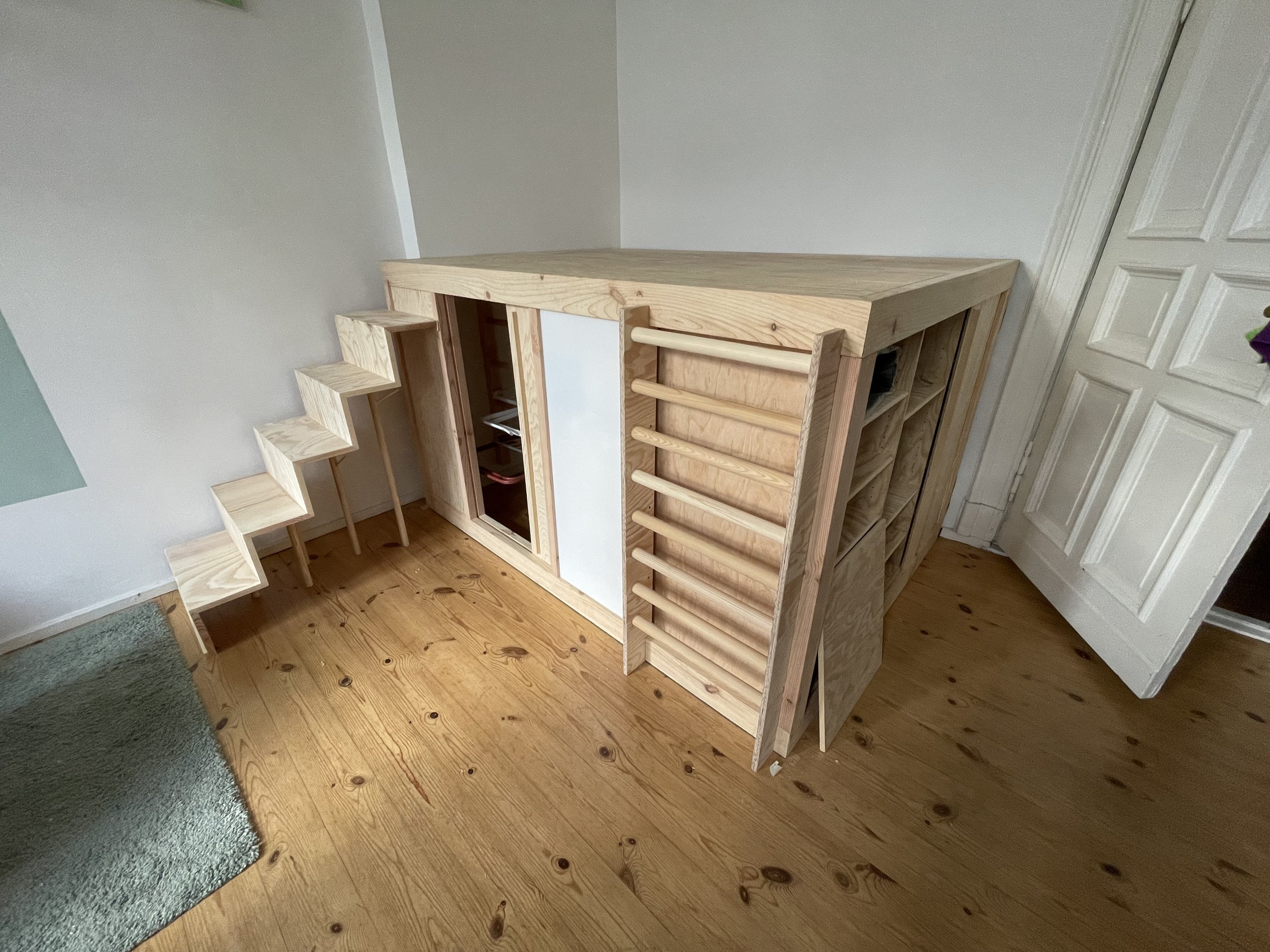 Ein Holzspielhaus für Kinder mit Leiter, Fenster und Verkleidung, auf Holzboden in einem Zimmer
