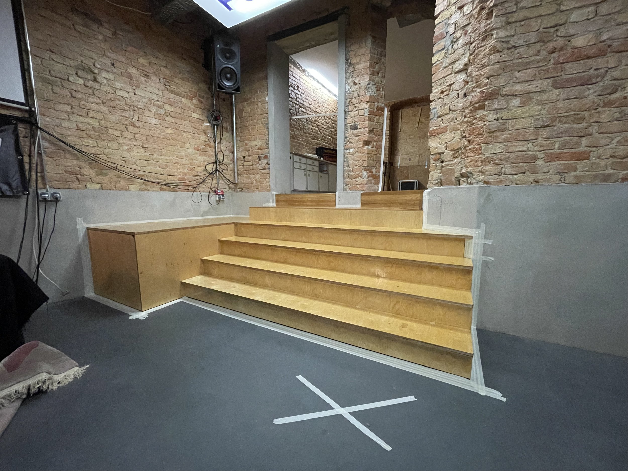 Innenraum mit roten Backsteinwänden und einer Holztreppe, die in einen anderen Raum führt, sowie Kabel und Lautsprecher an der Wand.