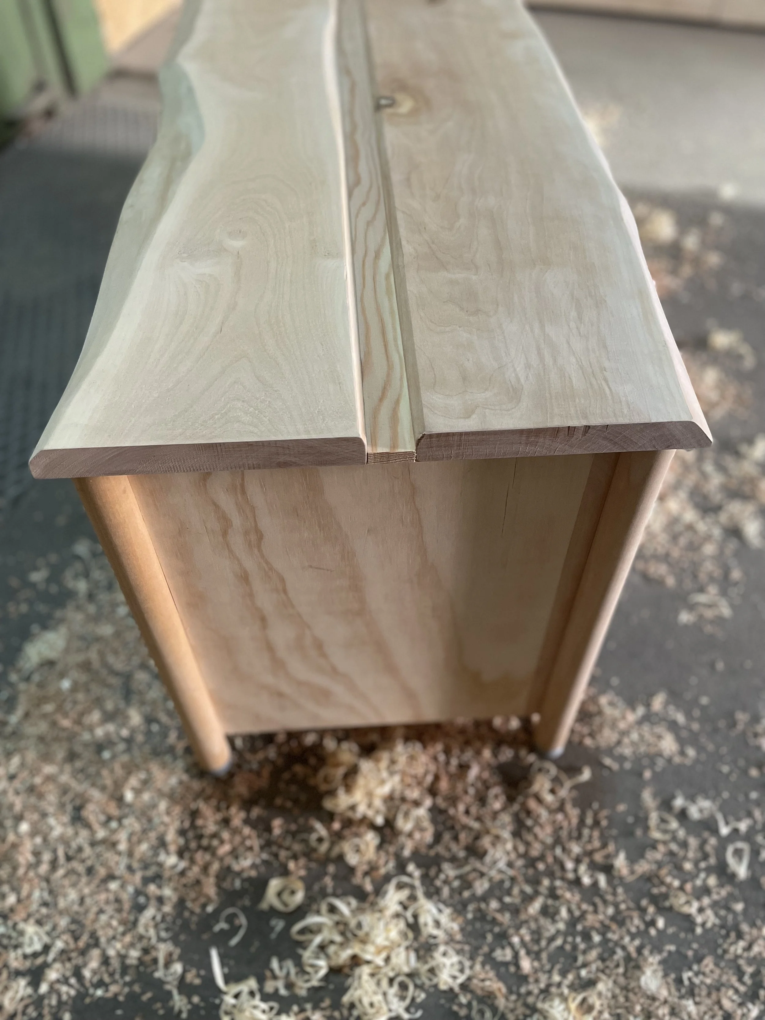 Unvollendeter Holzschrank aus hell lackiertem Holz mit sichtbarem Maserungen, Holzspäne liegen auf dem Boden.