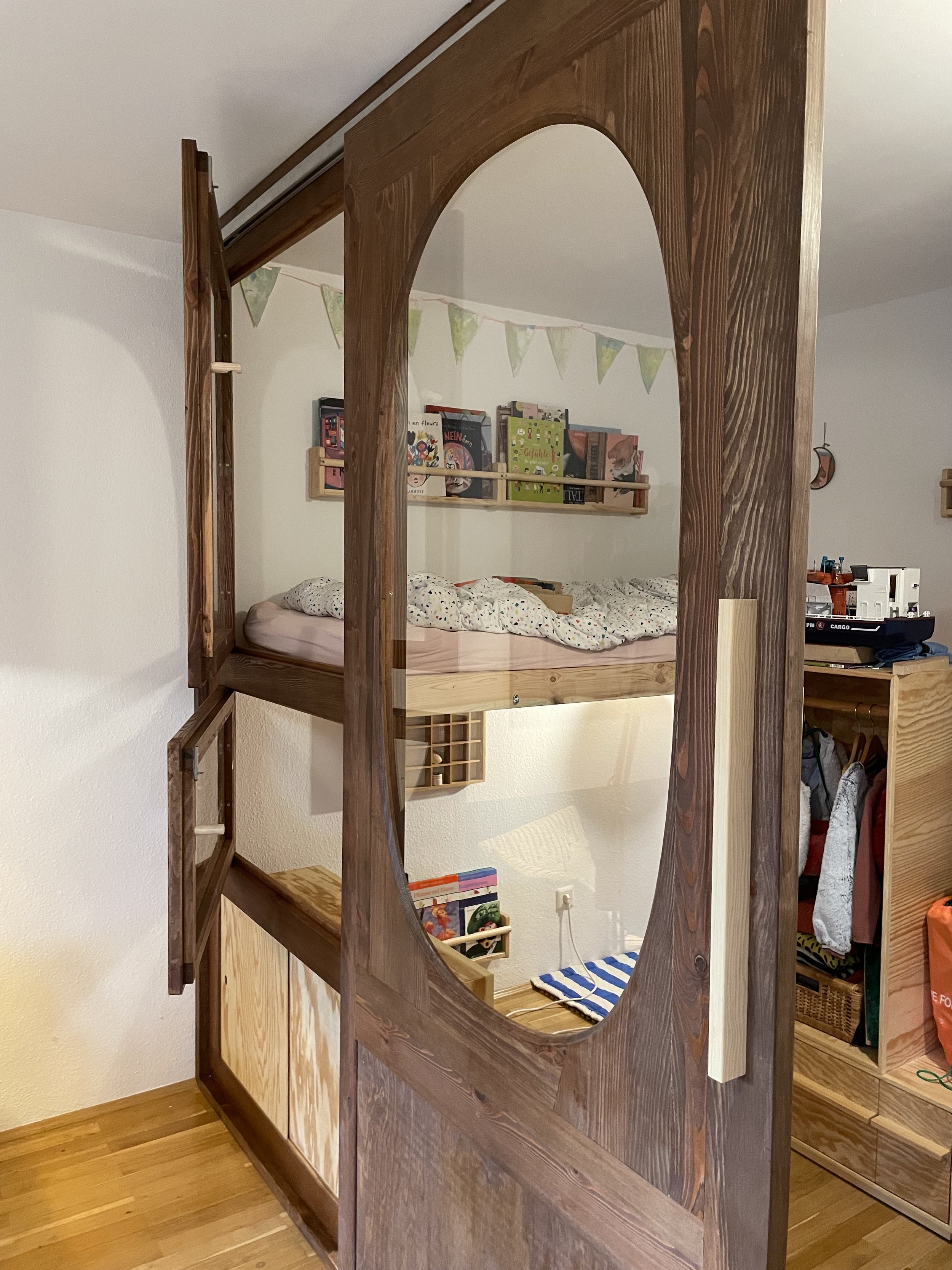 Blick durch eine hölzerne Raumtrenneröffnung auf ein Kinderbett auf Hochbett mit Bücherregal und Dekorationen im Kinderzimmer.