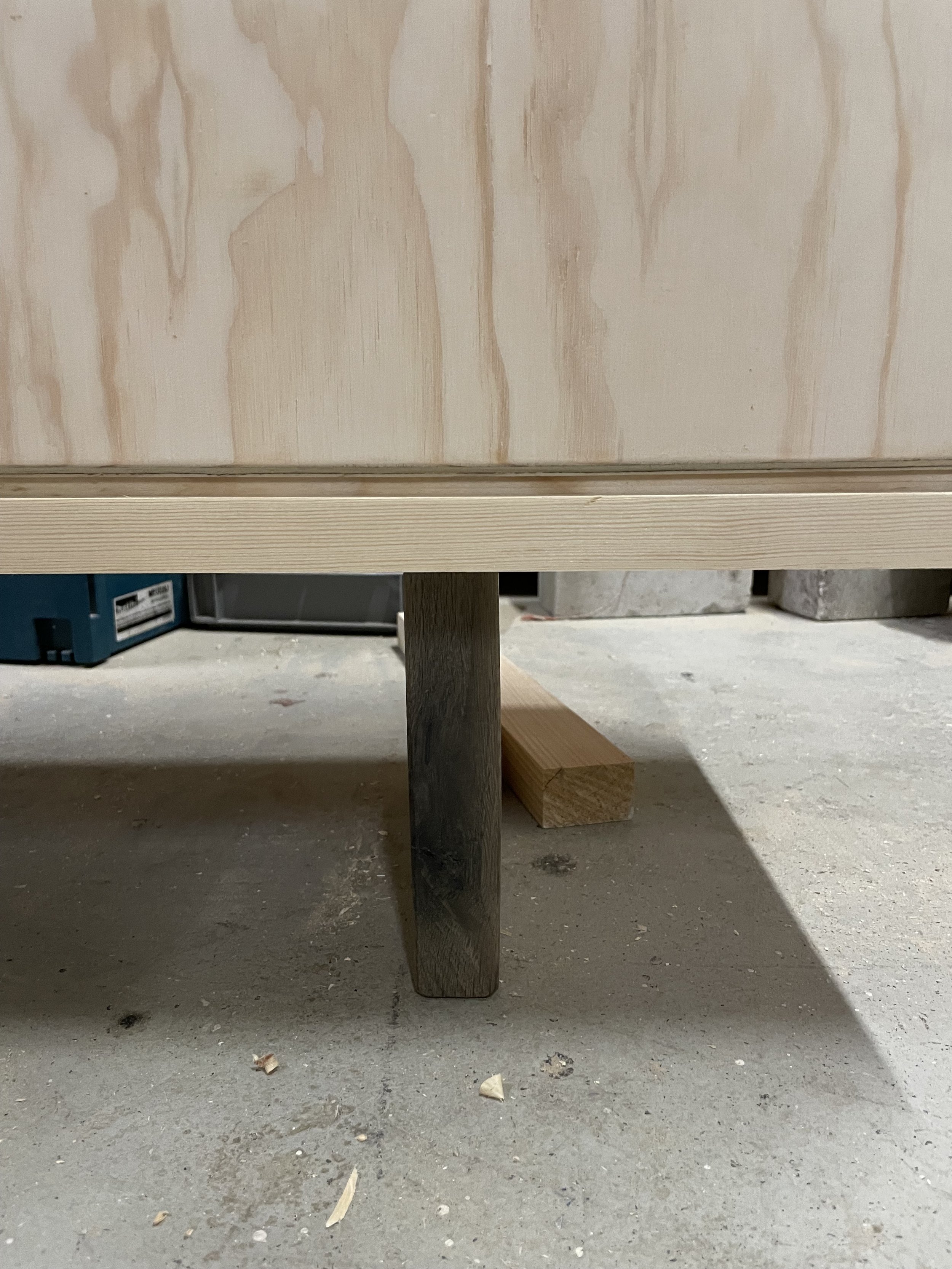 Unvollendeter Möbelrahmen auf einem Betonboden, mit Holzbeinen und Holzplatten. Im Hintergrund Werkstattumgebung sichtbar.