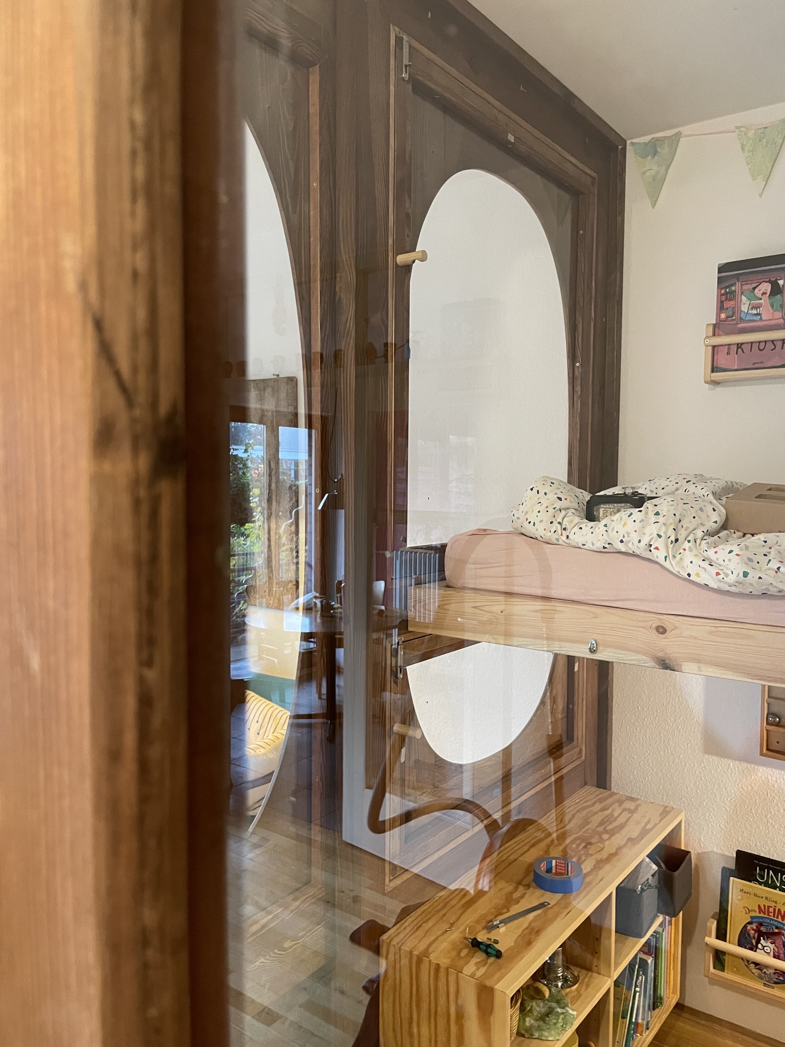 Blick durch eine Glasscheibe in ein Zimmer mit einem Holzregal und einem Bett, auf dem Decke und ein Wecker liegen, hinter dem Glas sind bunte Wimpel und Bücher sichtbar.