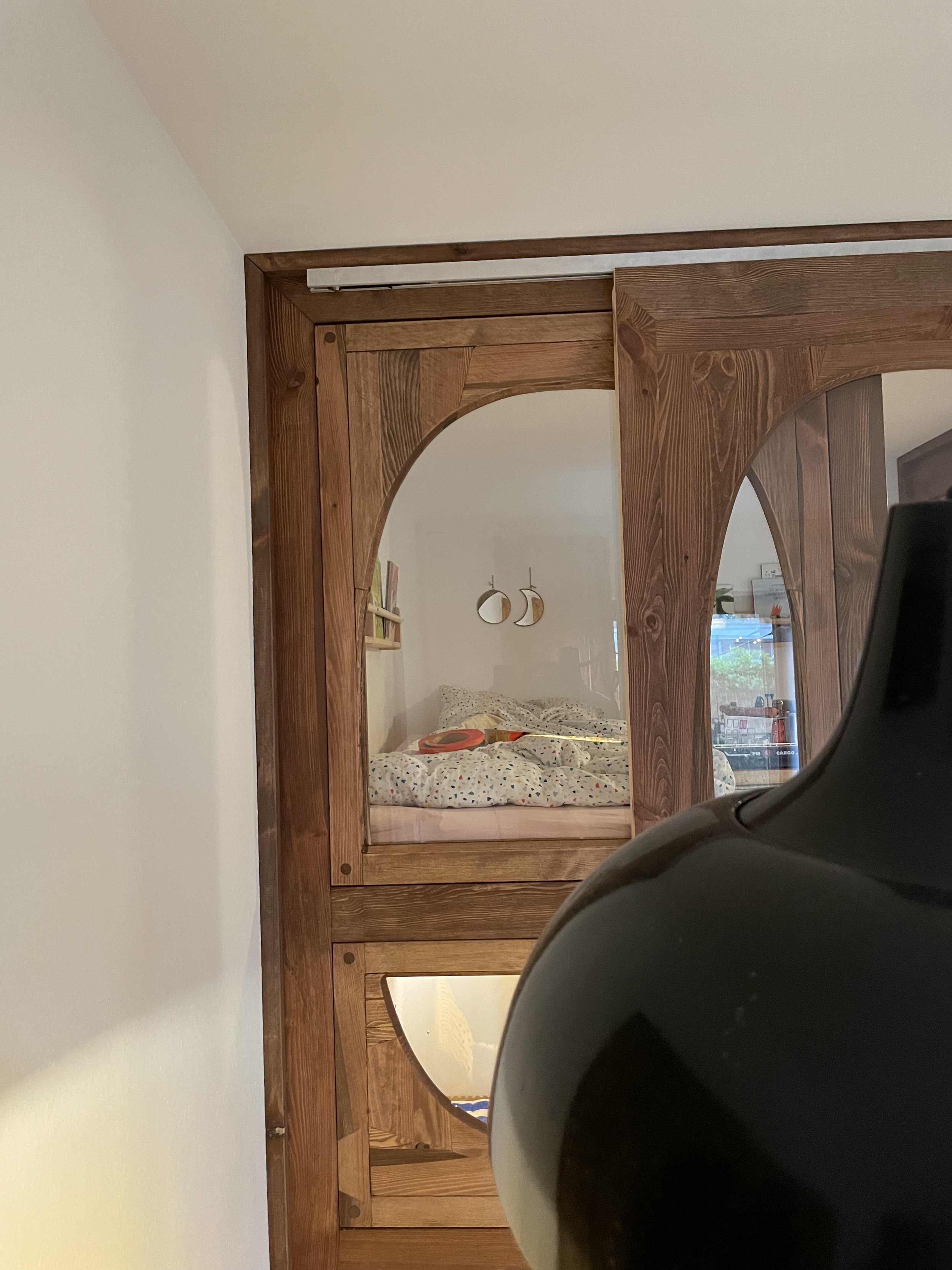Blick in ein Schlafzimmer durch eine halb offene Holz-Schiebetür, reflektiert im Spiegel auf der Tür. Im Schlafzimmer sind zwei runde Wandspiegel und ein Bett mit buntem Bettbezug sichtbar.