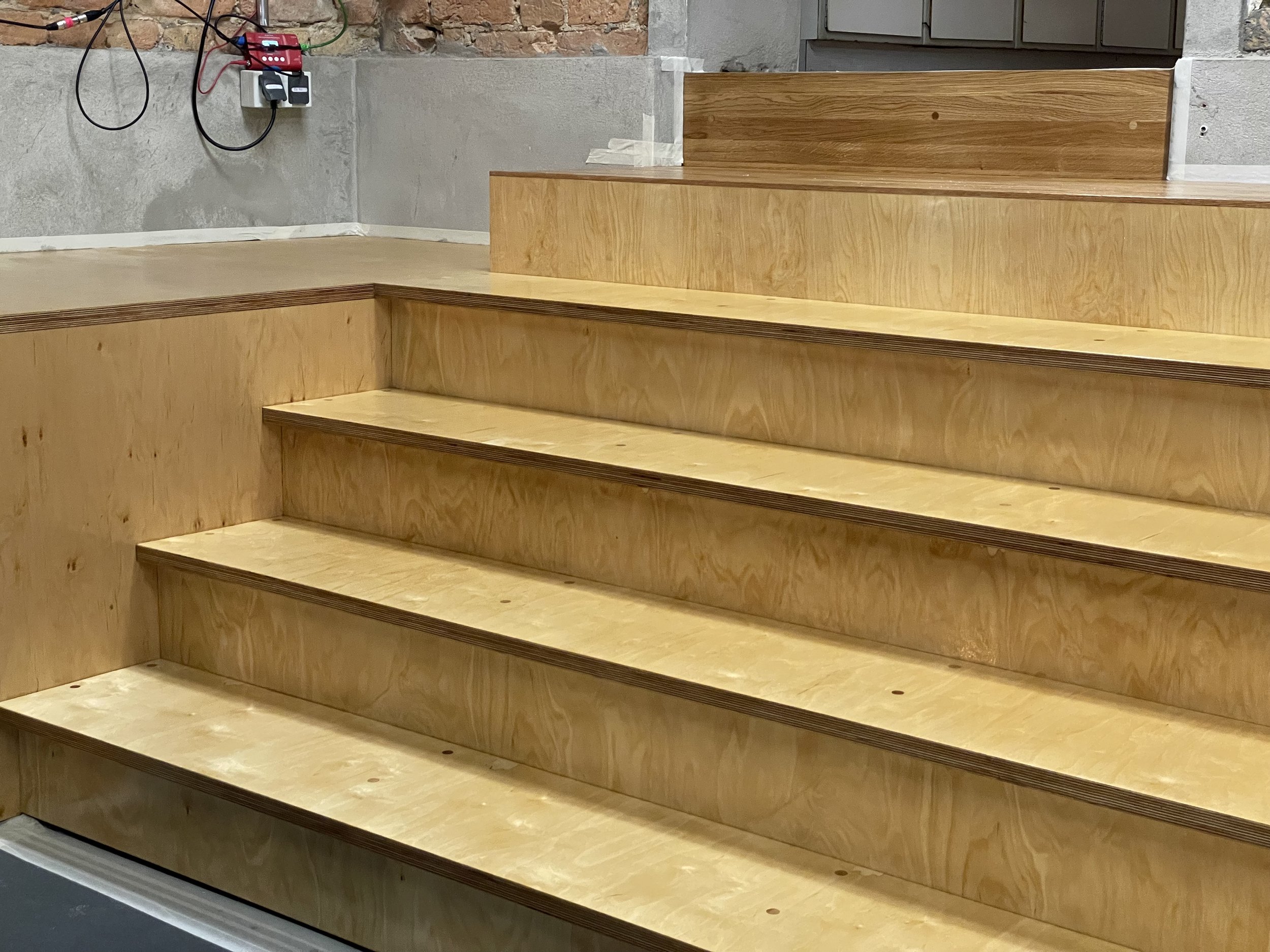 Holztreppe mit vier Stufen, im Hintergrund eine Wand aus Ziegeln mit elektrischer Steuerung und Kabel.