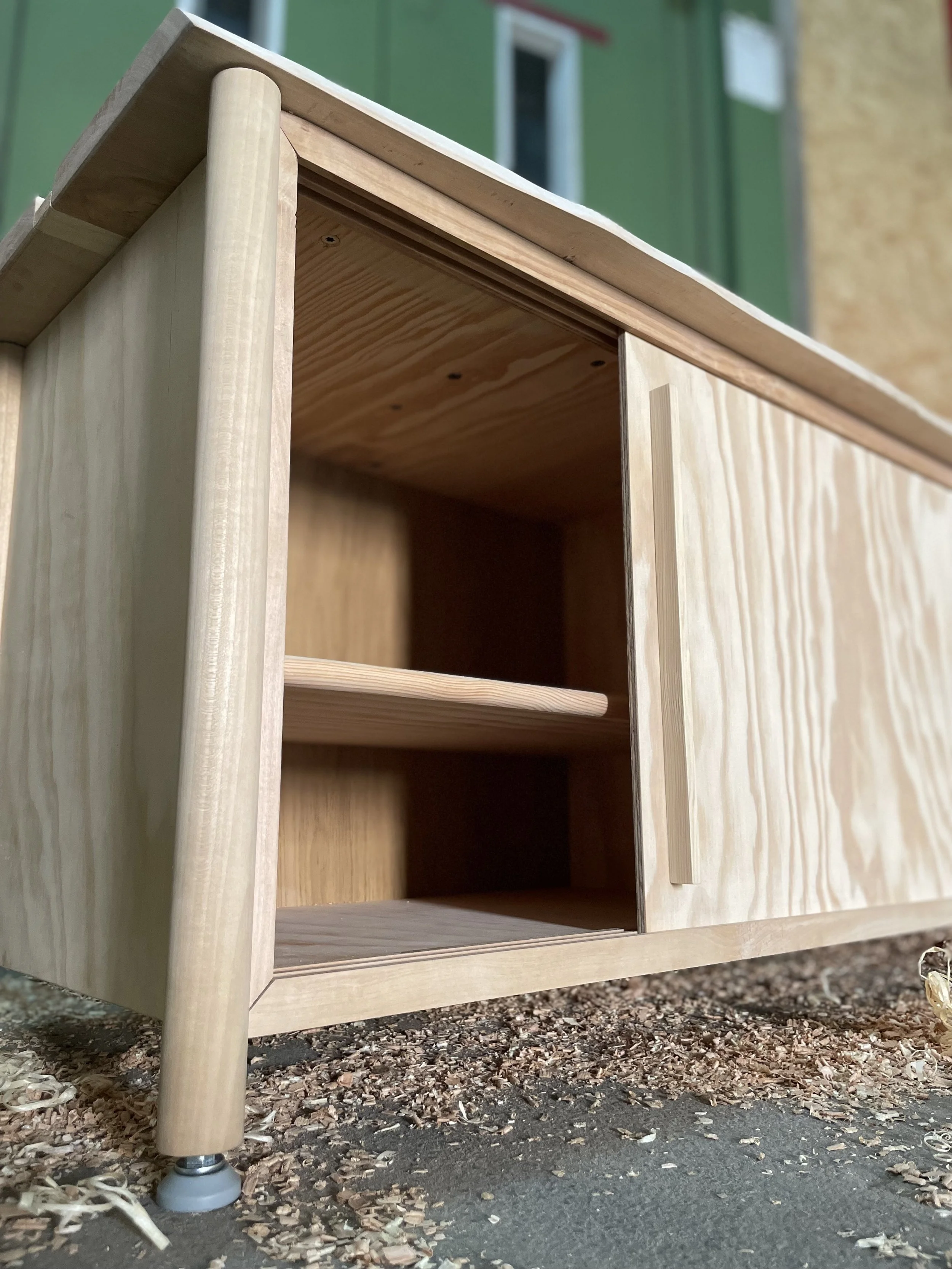 Nahaufnahme eines kleinen, handgefertigten Möbelstücks aus Holz, vermutlich eine kleine Schublade oder ein Regal, auf einer Oberfläche mit Holzspänen und Staub, vor einem grün lackierten Gebäude mit Fenstern.