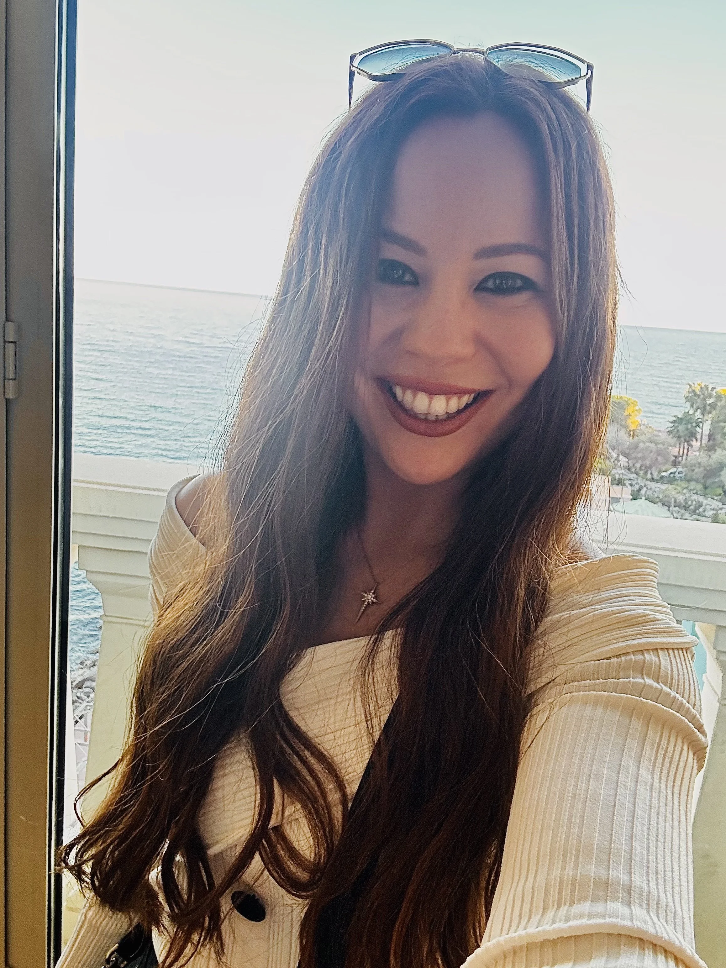 A woman with long wavy brown hair taking a selfie indoors near a window overlooking the ocean, wearing sunglasses on her head, a beige top, a necklace with a star pendant, and smiling.