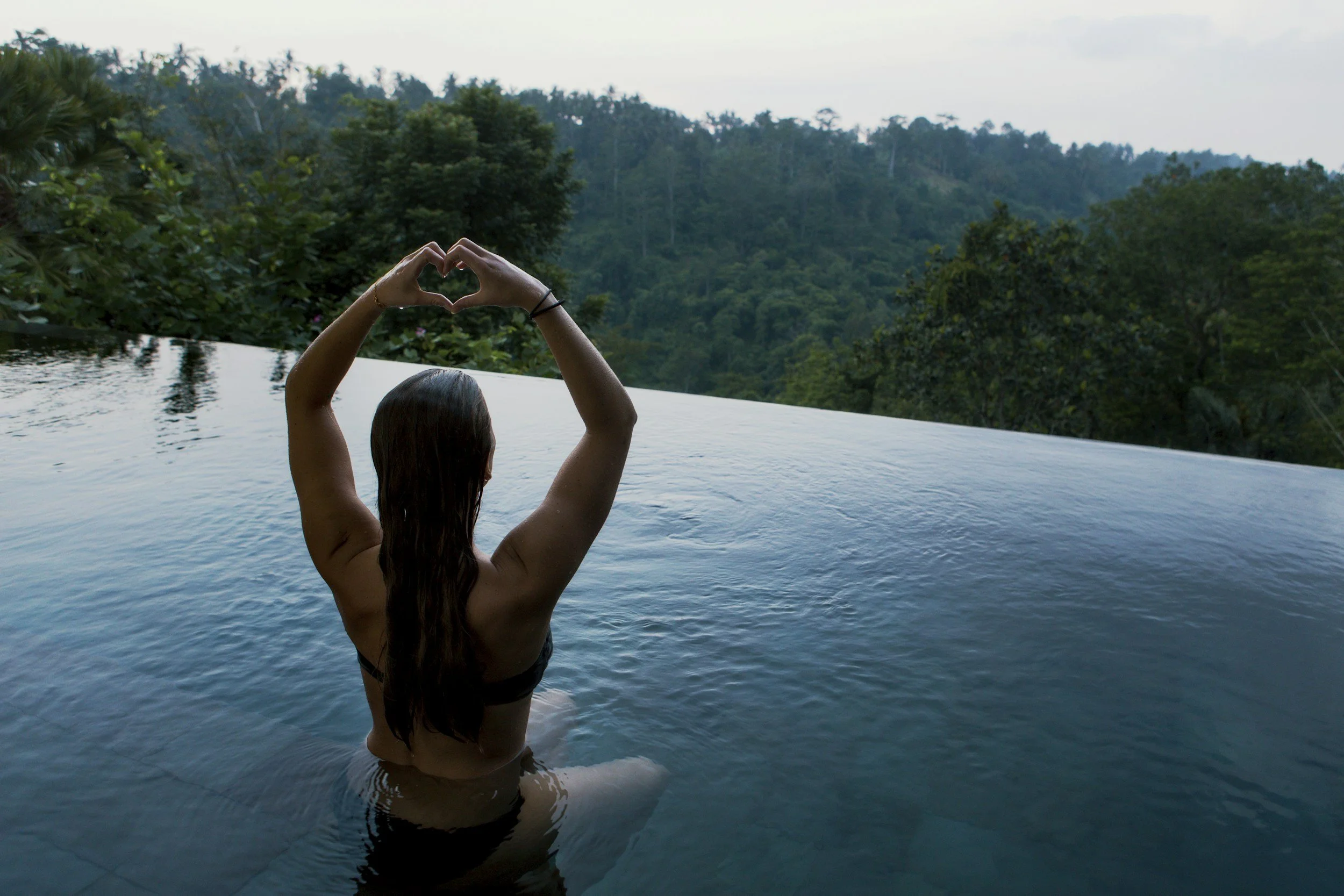 A woman with long dark hair swimming in an infinity pool overlooking a lush green forest and hills, making a heart shape with her hands.