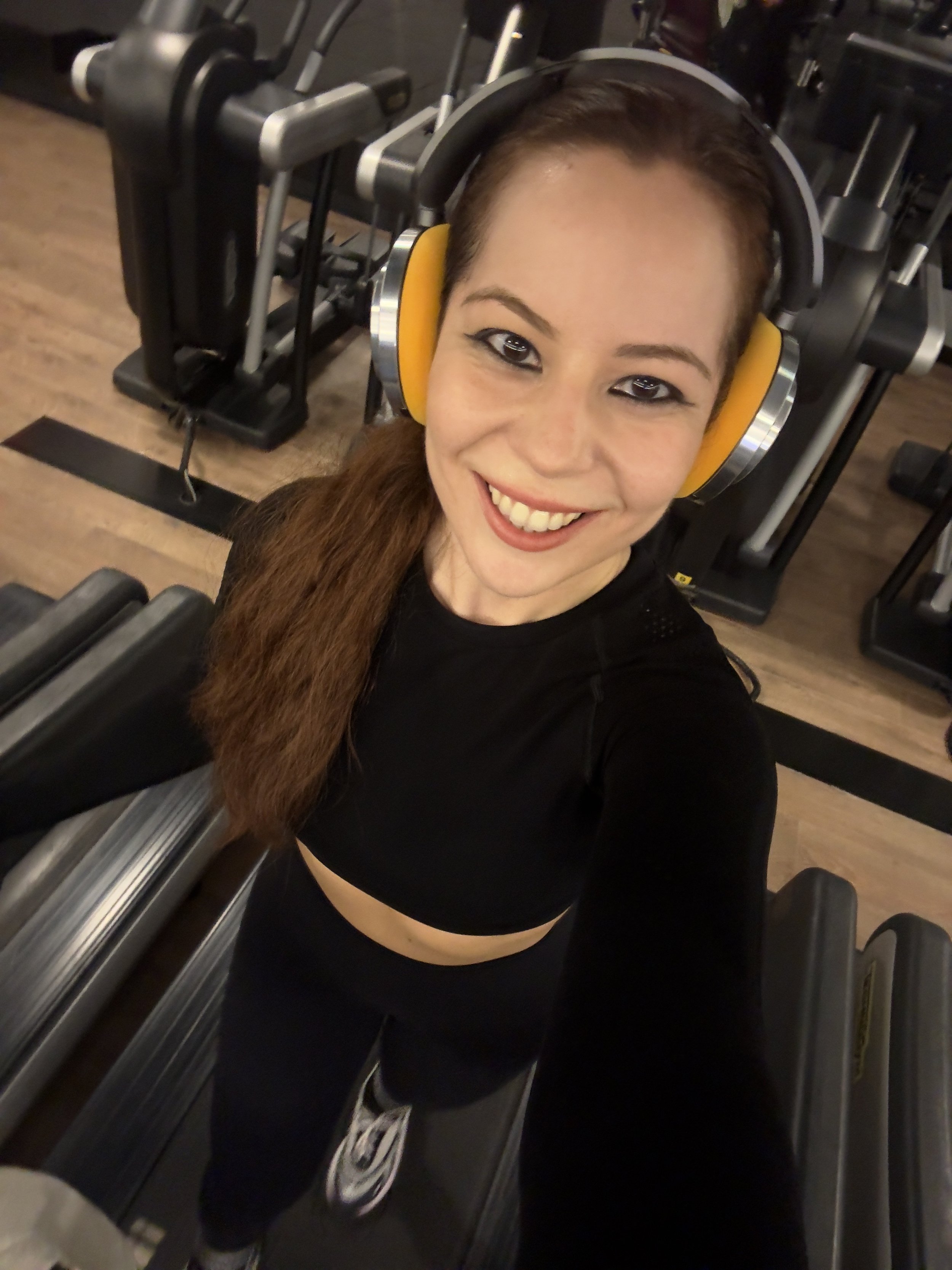 Woman in workout clothes smiling at the camera at the gym, wearing yellow ear protection, standing on an exercise treadmill.
