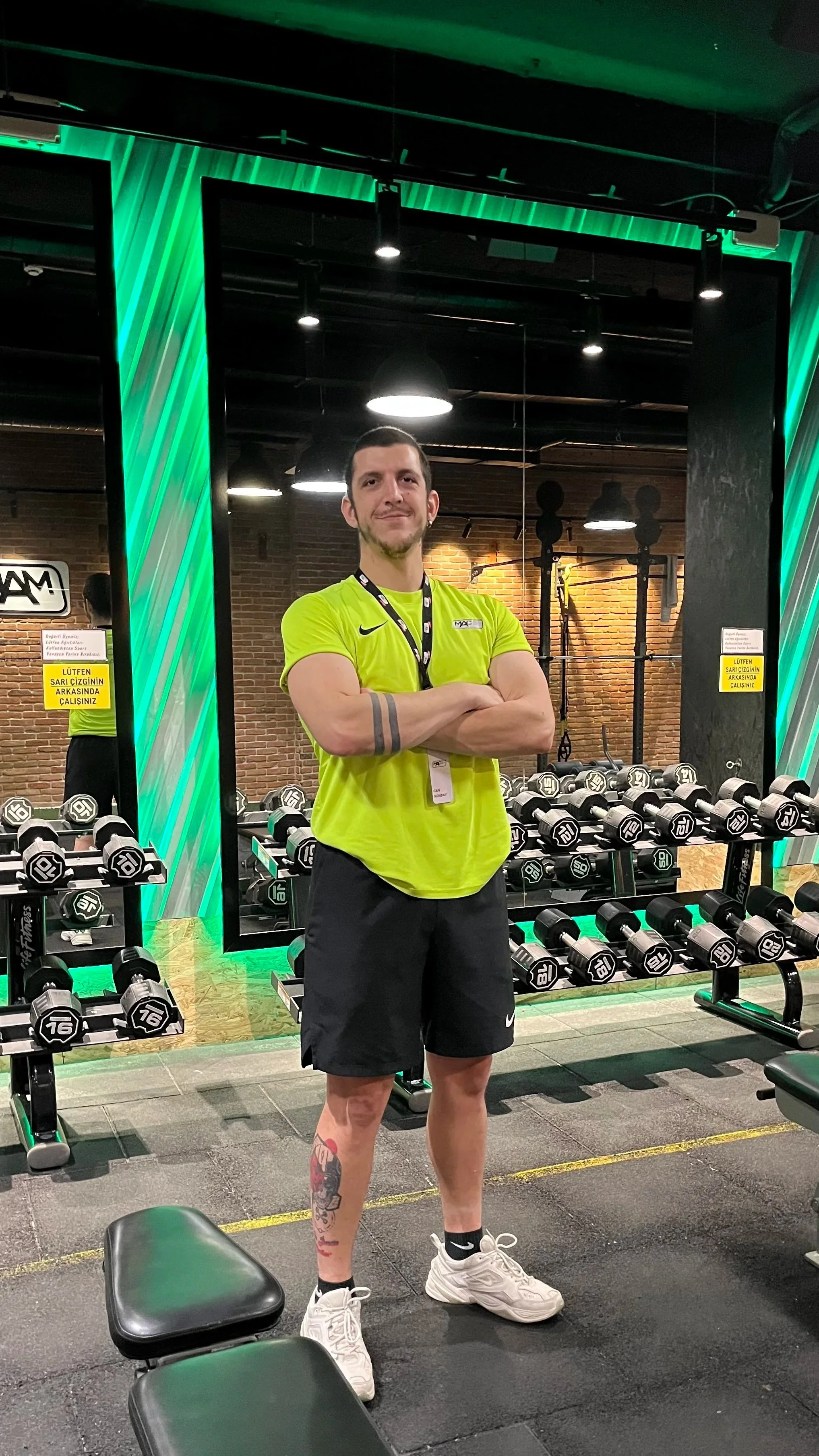 A man standing in a gym in front of a mirror with dumbbells and workout equipment, wearing a neon yellow shirt, black shorts, and white sneakers, with arms crossed and a slight smile.
