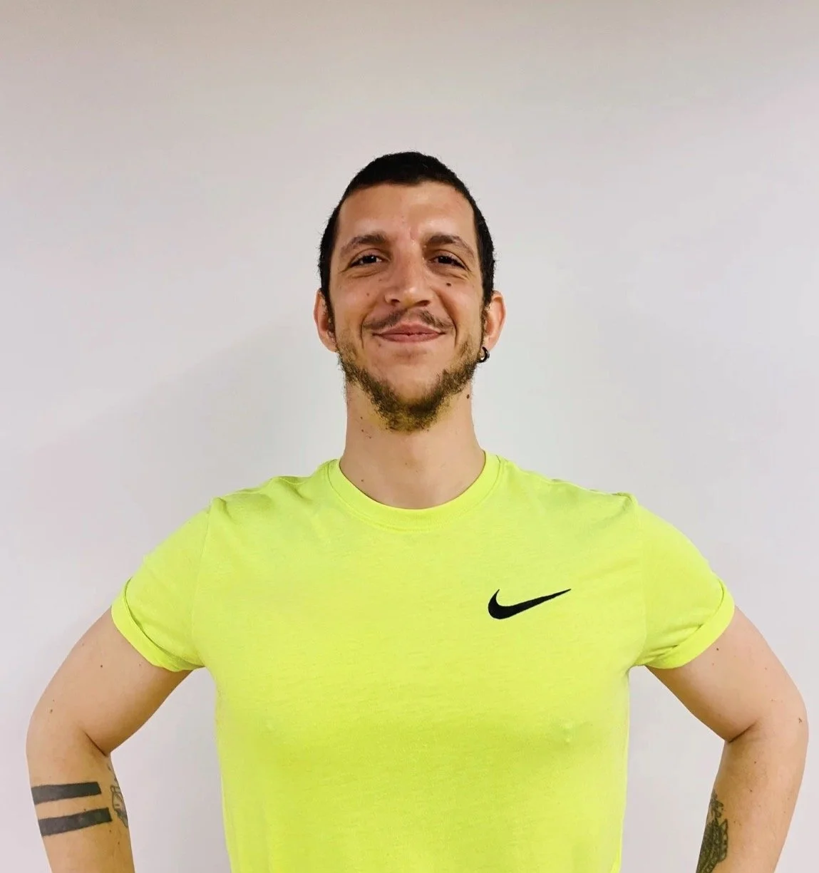 A young man with short dark hair, a goatee, and earrings in both ears, standing against a plain white wall. He is smiling and wearing a bright yellow Nike T-shirt.