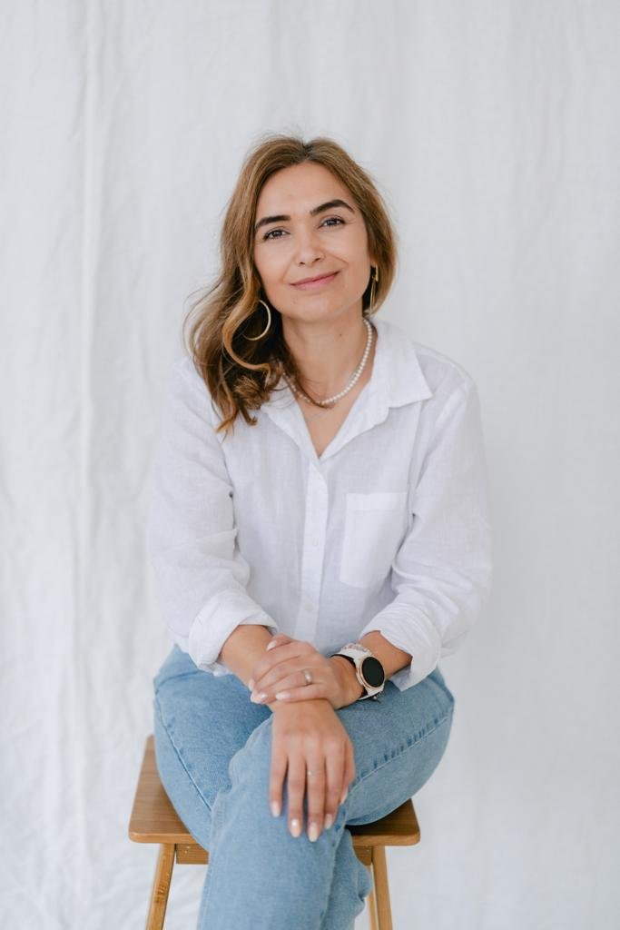 Person sitting on a wooden stool in a white shirt and blue jeans, with a smartwatch and hoop earrings. Background is a plain, light-colored curtain.