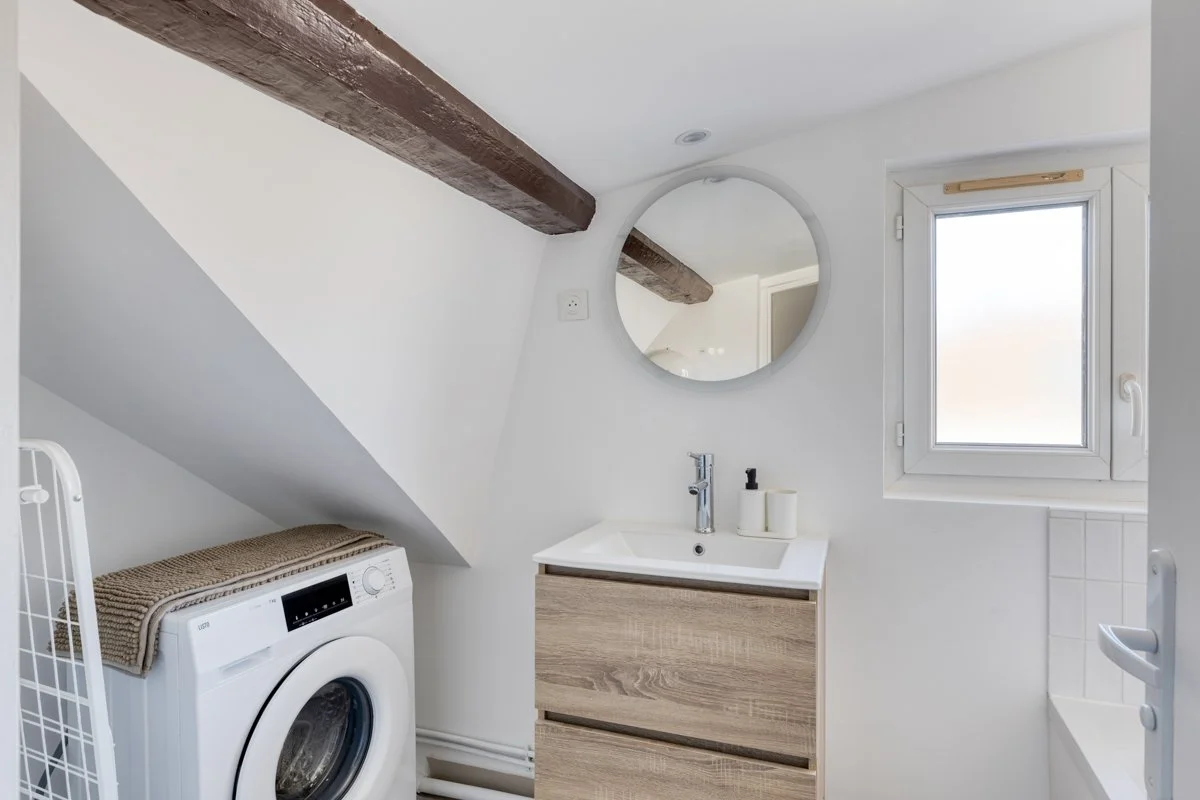 Petite salle de bain avec lavabo, miroir rond, fenêtre, et machine à laver sous un plafond incliné avec poutre en bois.